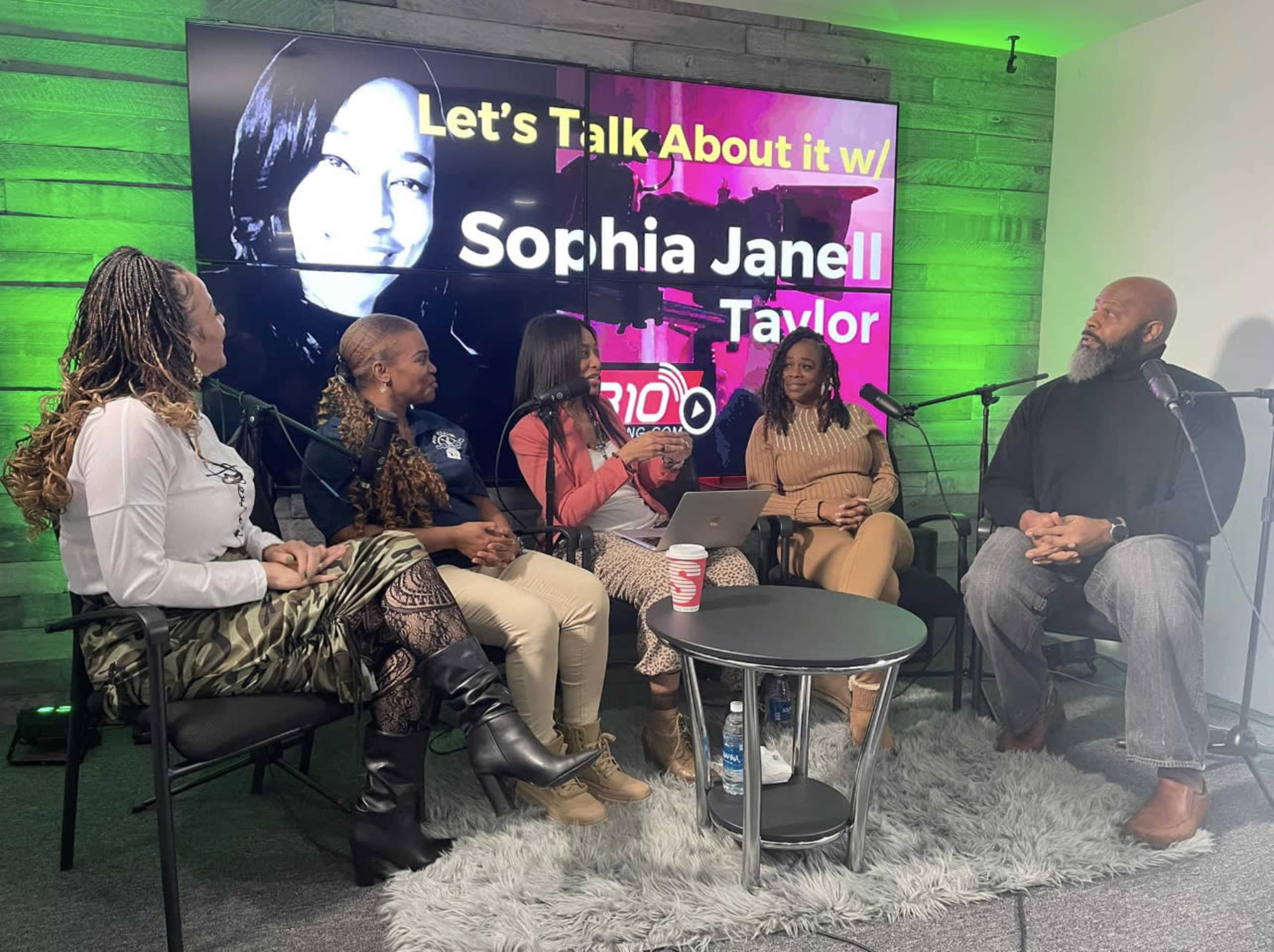 A group of five people sits around a table with microphones, discussing a topic on a set with a large screen displaying the words "Let's Talk About It w/ Sophia Janell Taylor."
