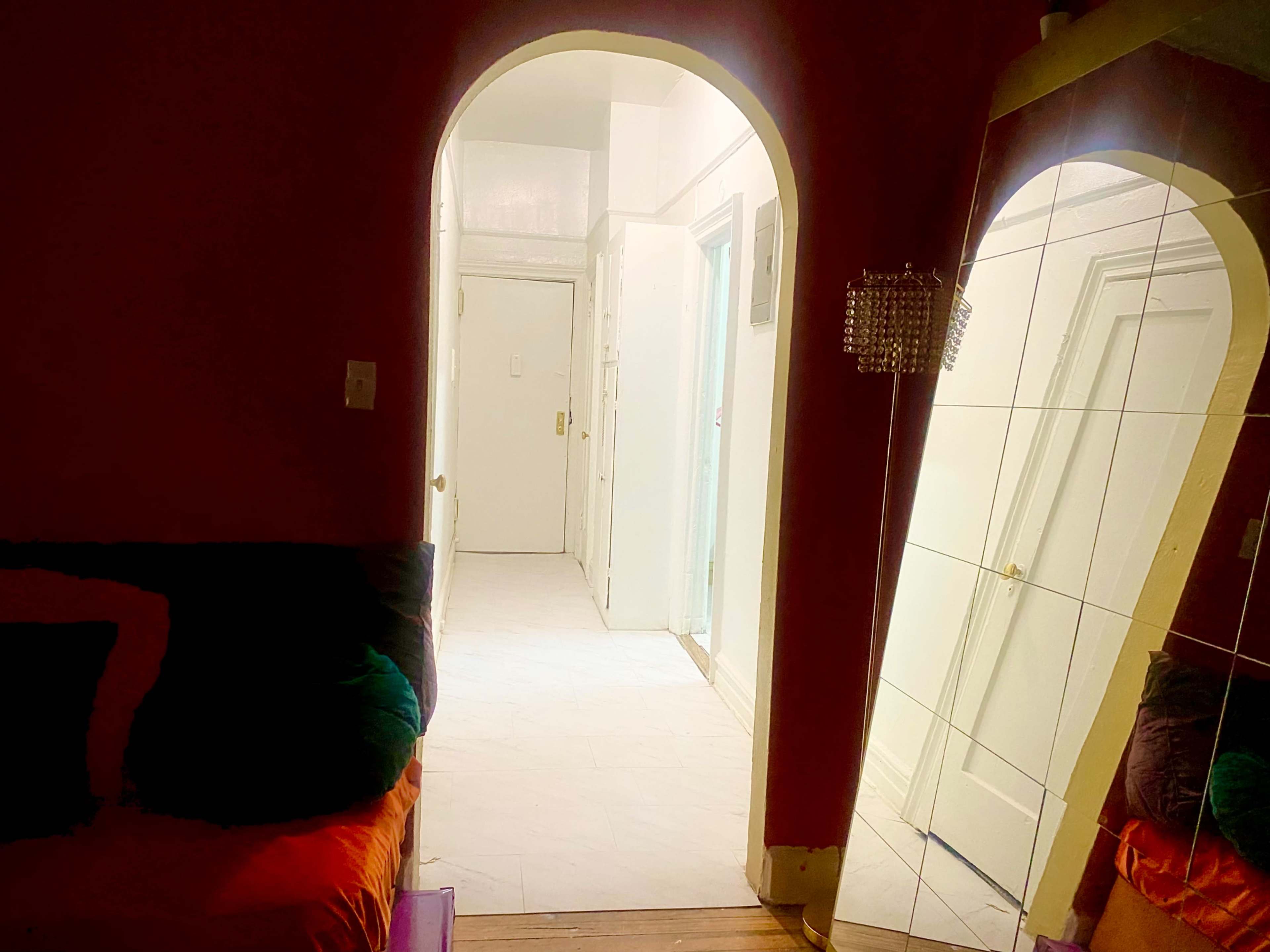 A dimly lit hallway with an archway leading to a white-tiled area and a mirrored wall reflecting a colorful couch.