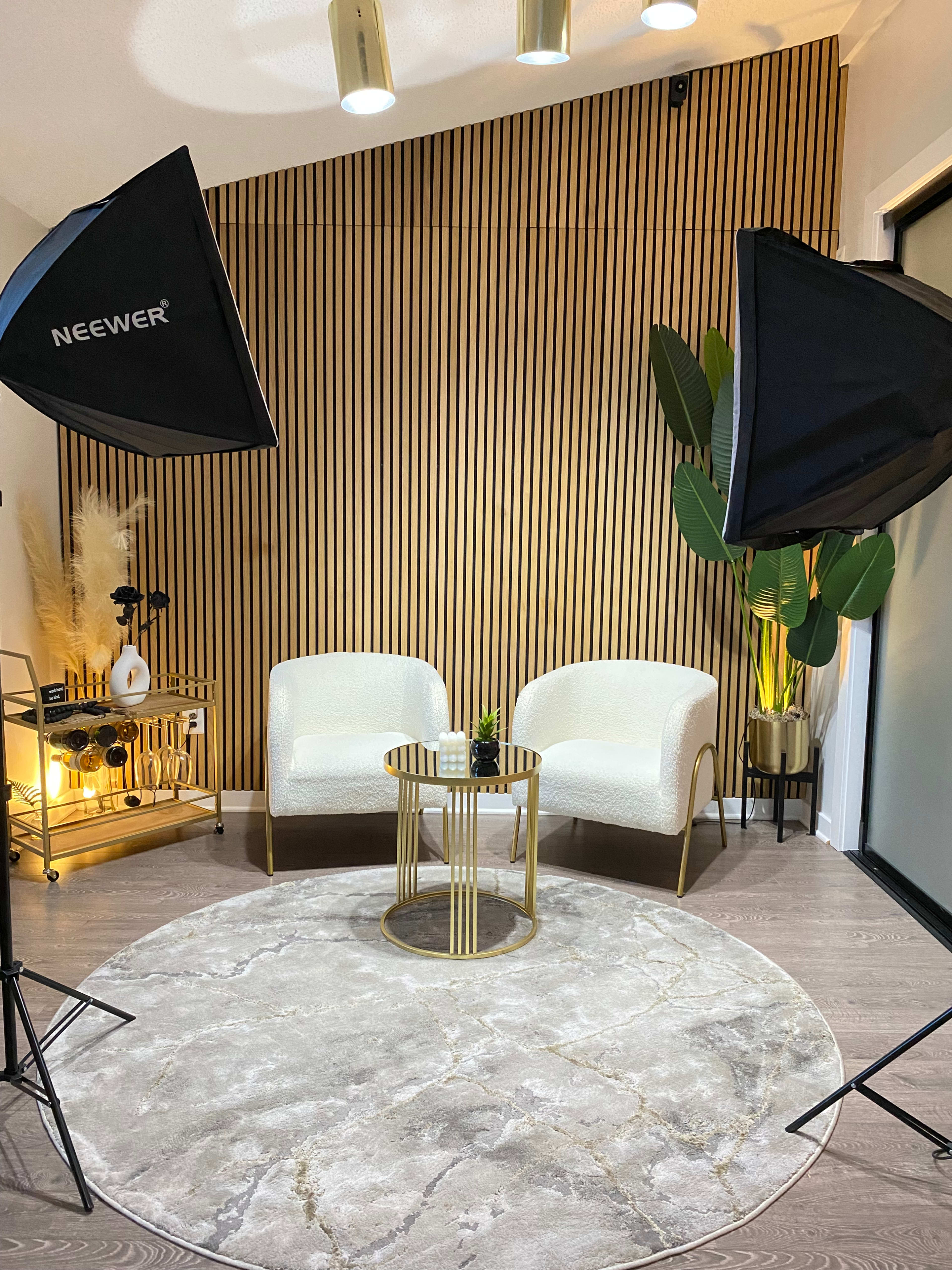 The image shows a modern interior setup featuring two white chairs, a small gold and glass table, a round gray rug, and two photography lights, all arranged against a striped wood accent wall.