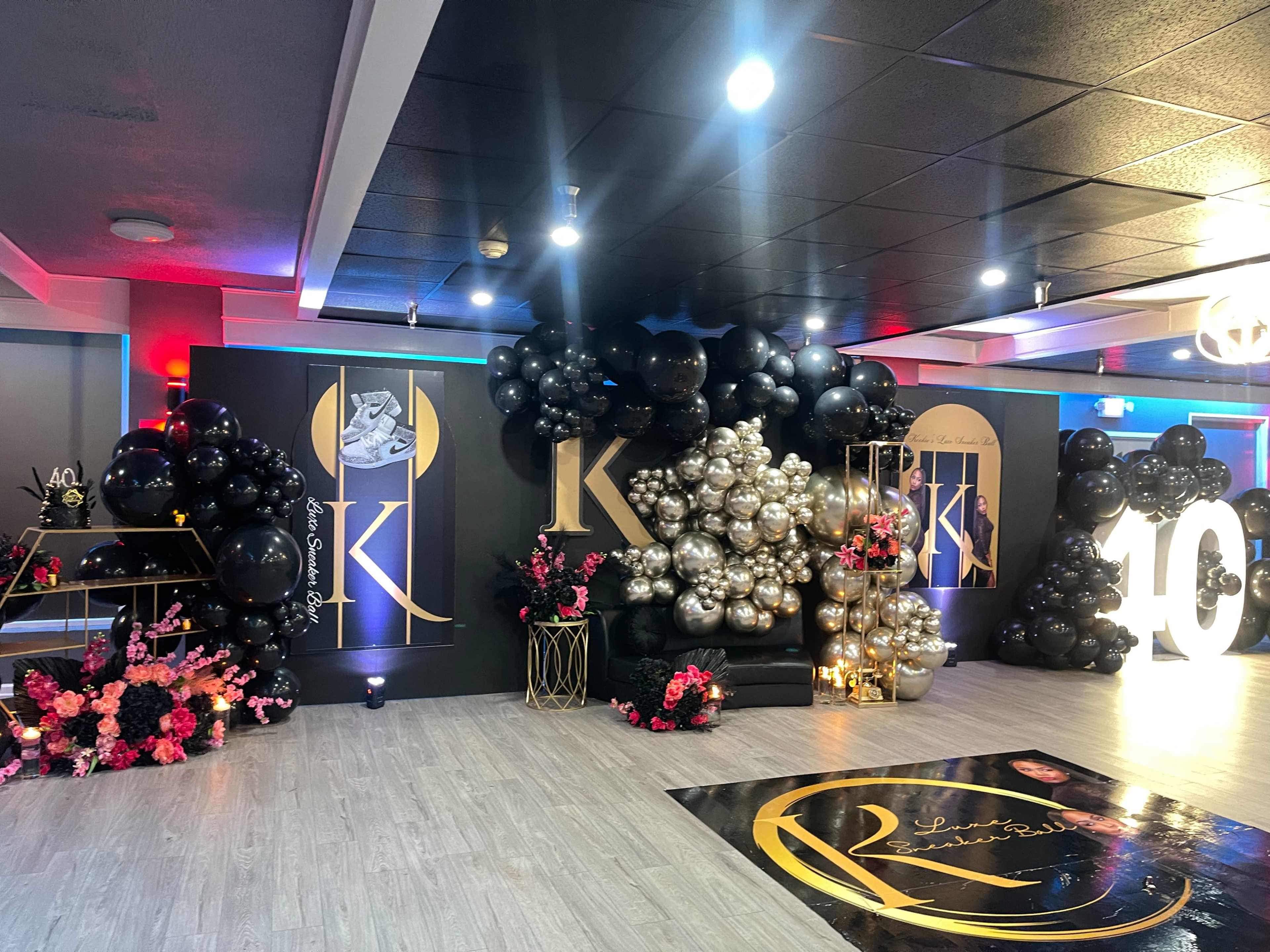 The image shows a decorated event space with black and gold balloons, floral arrangements, and large display numbers.