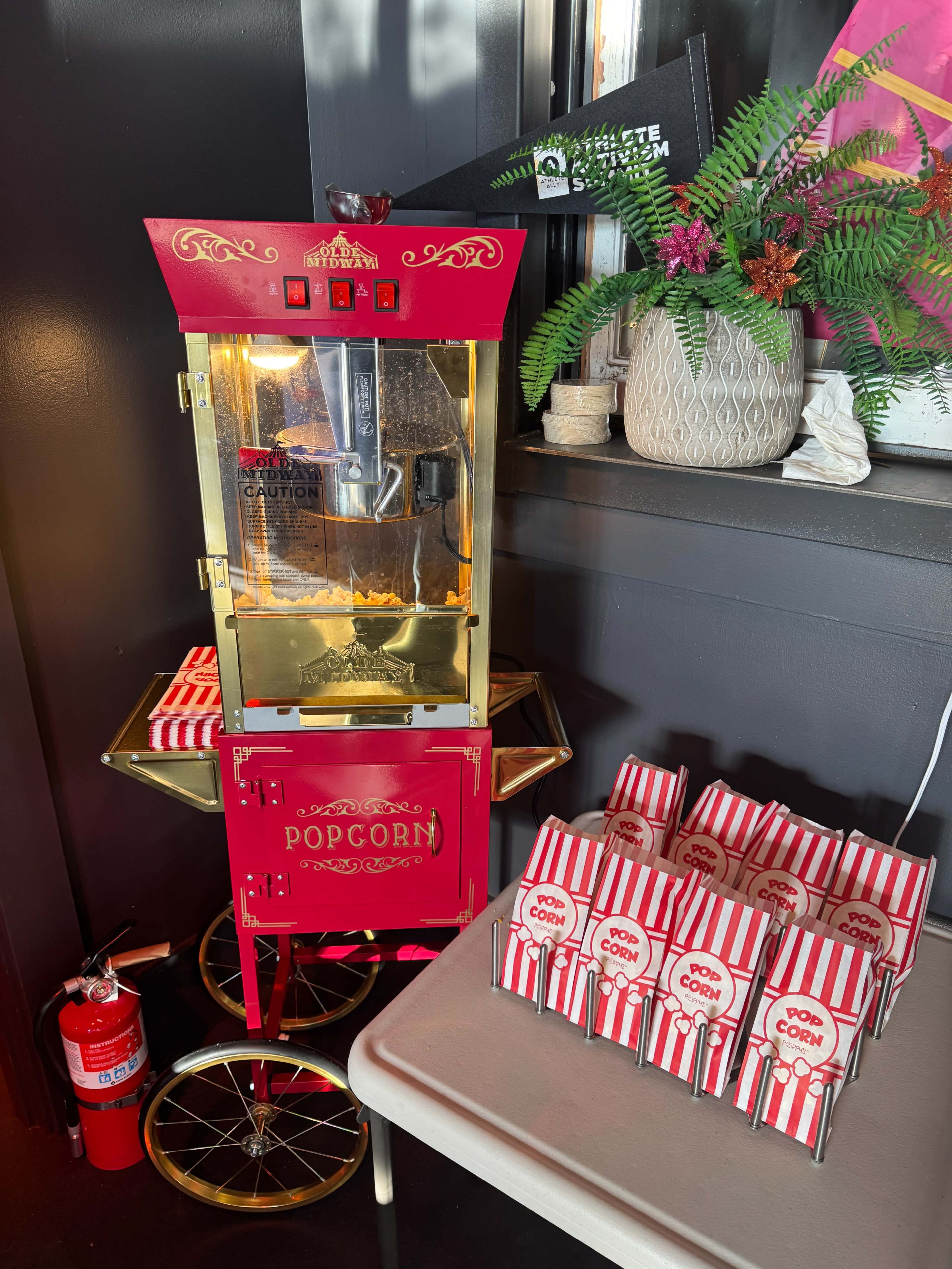A vintage-style red popcorn machine stands next to a table stacked with striped popcorn boxes in a brightly lit space.