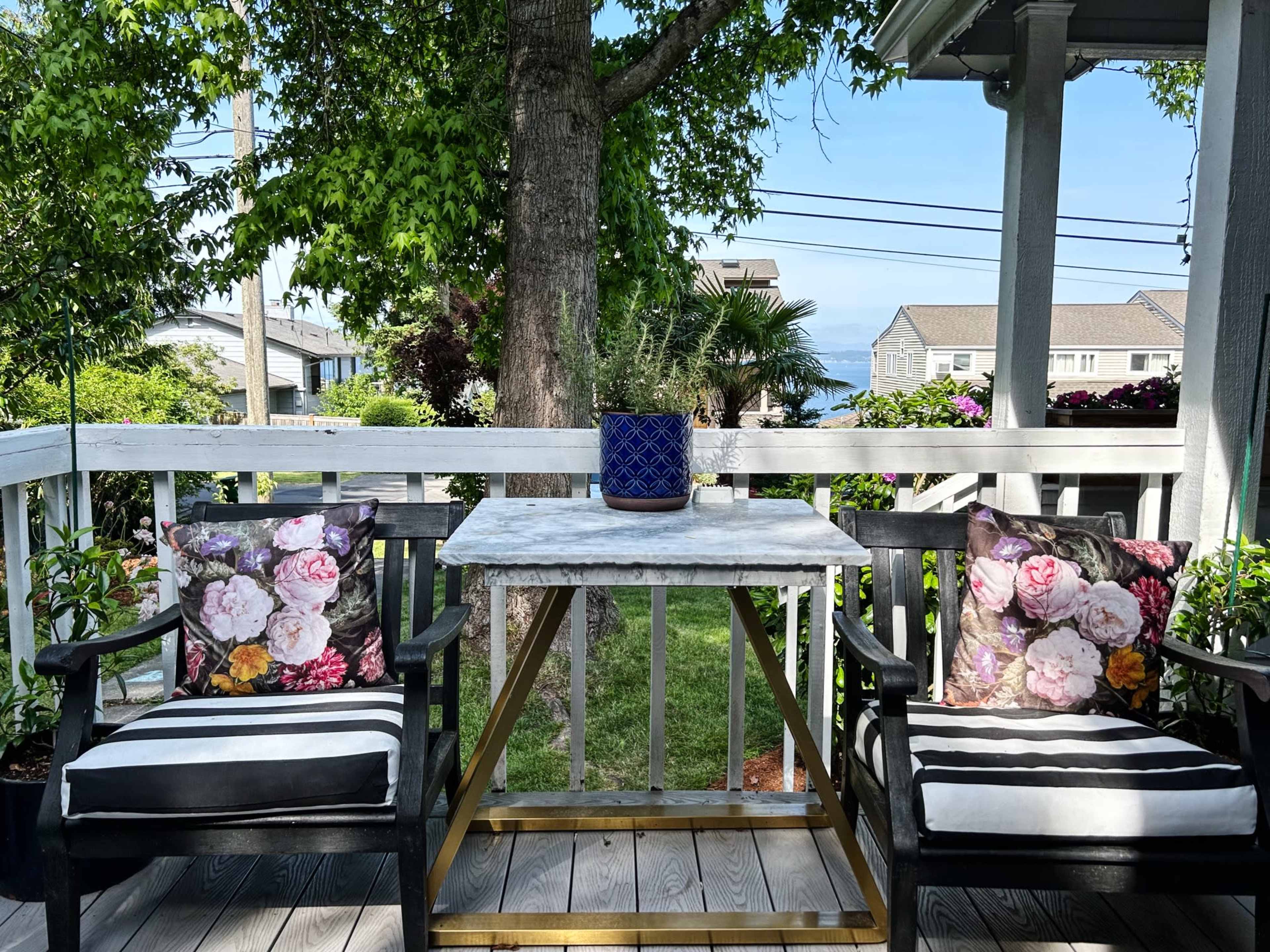 A porch with two striped chairs and a marble-topped table, surrounded by greenery and a view of water in the background.