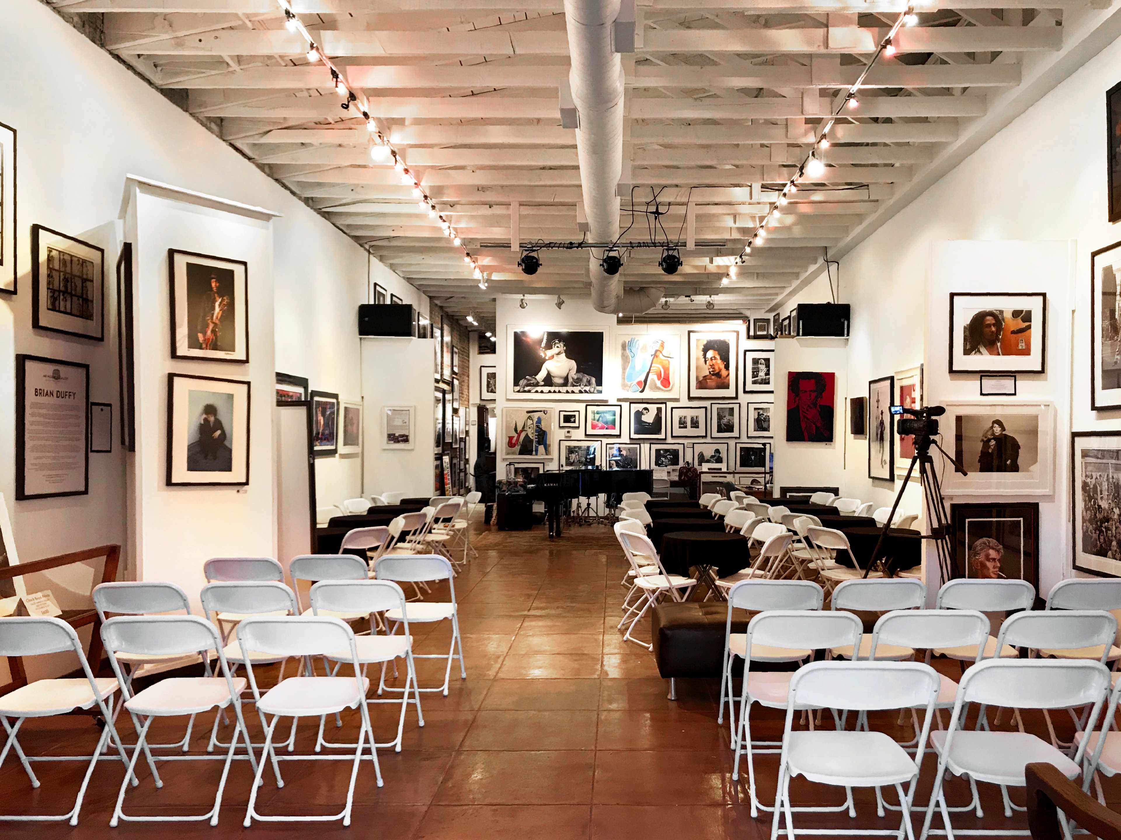 A gallery interior with white folding chairs arranged for an event, surrounded by framed photographs on the walls and a piano at the back.