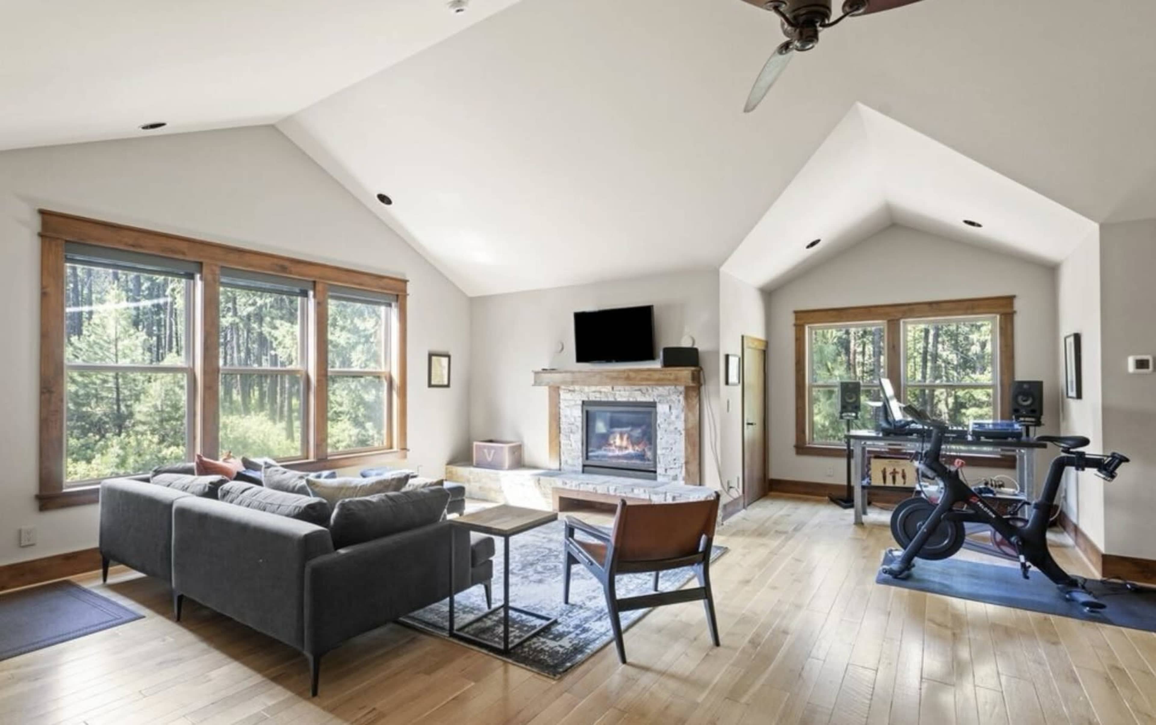 The living room features a fireplace, large windows showcasing trees outside, a seating area, and an exercise bike.