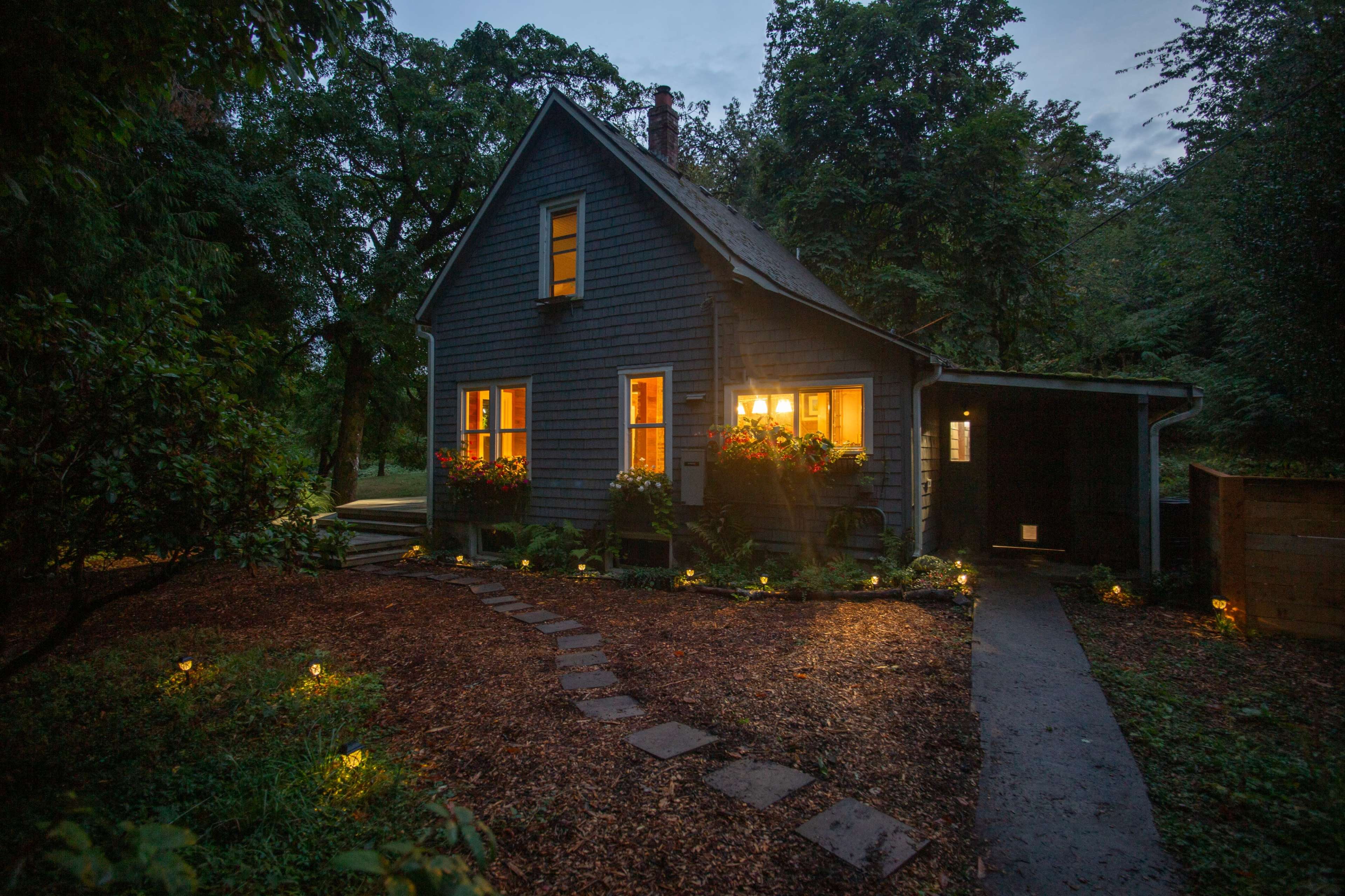 A two-story, dark blue house with lit windows and a stone pathway is surrounded by trees in a dimly lit evening setting.