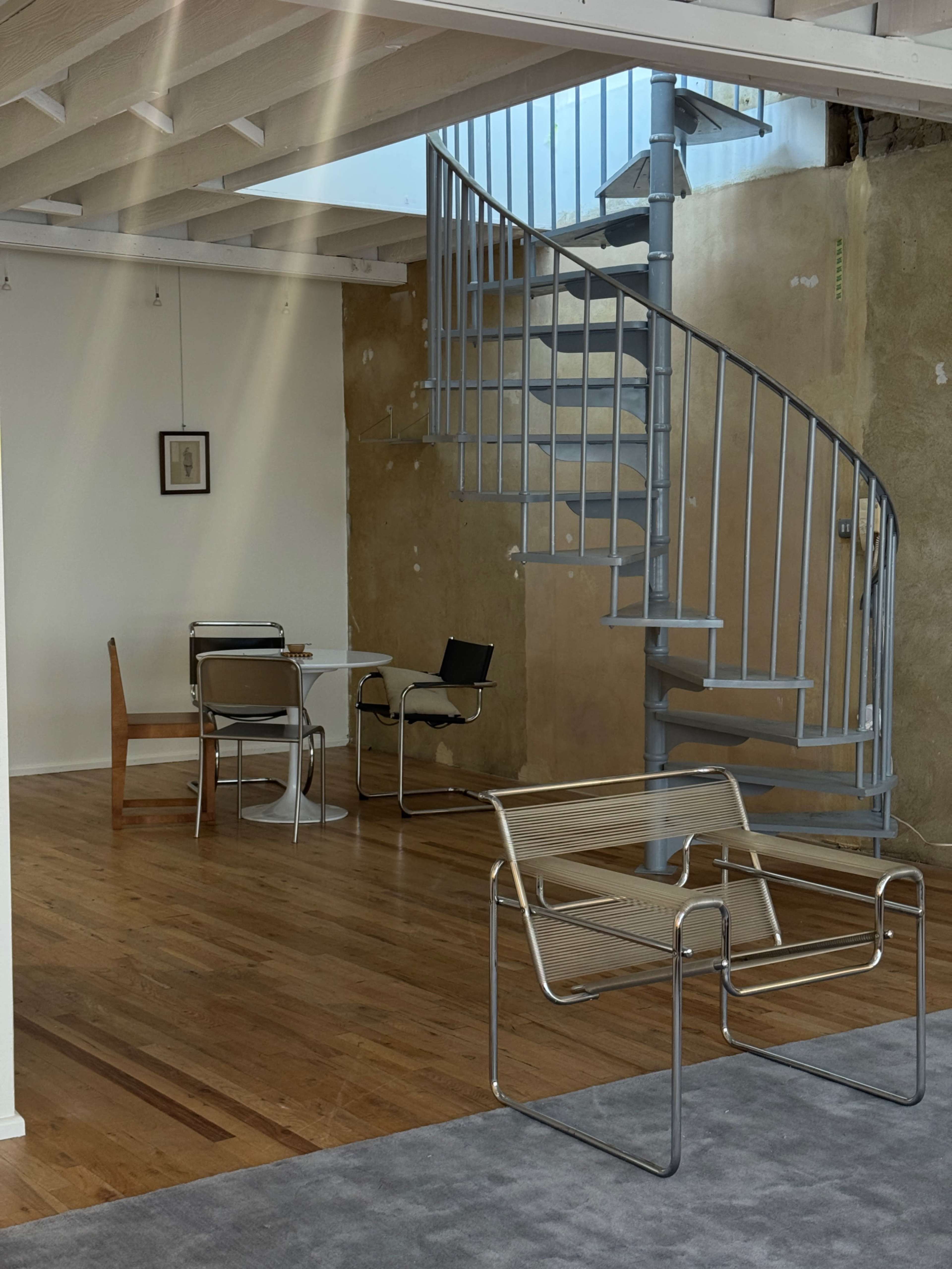 The image depicts a modern interior space featuring a spiral staircase, a small dining table with chairs, and minimalist furniture on wooden flooring.
