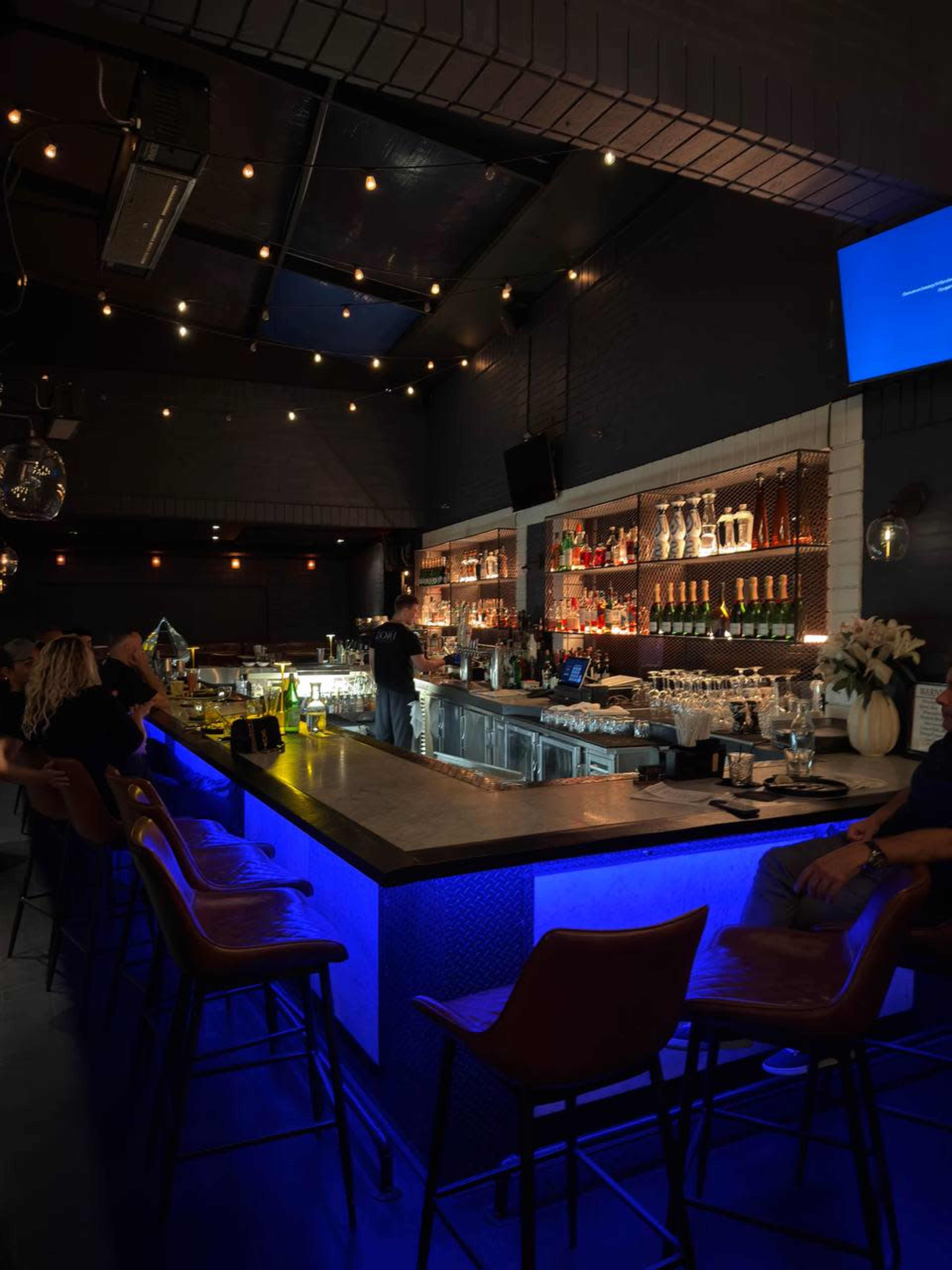 The image shows a modern bar interior with illuminated shelving displaying bottles, a bartender preparing drinks, and patrons seated at the bar.