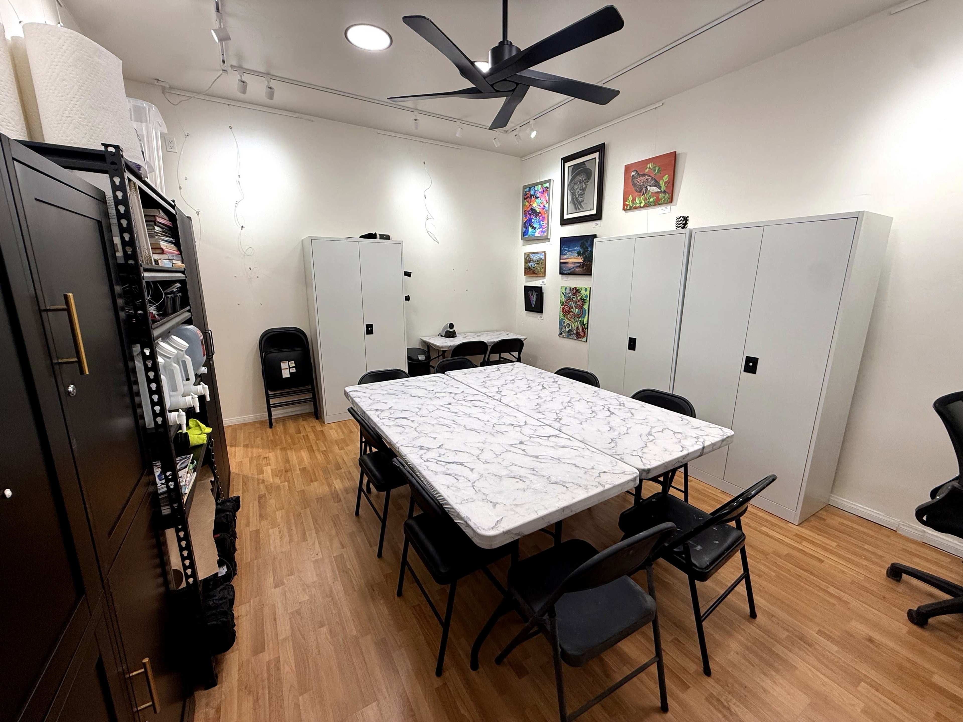 The image shows a well-lit room with a large table surrounded by black chairs, storage cabinets along the walls, and various artworks displayed.