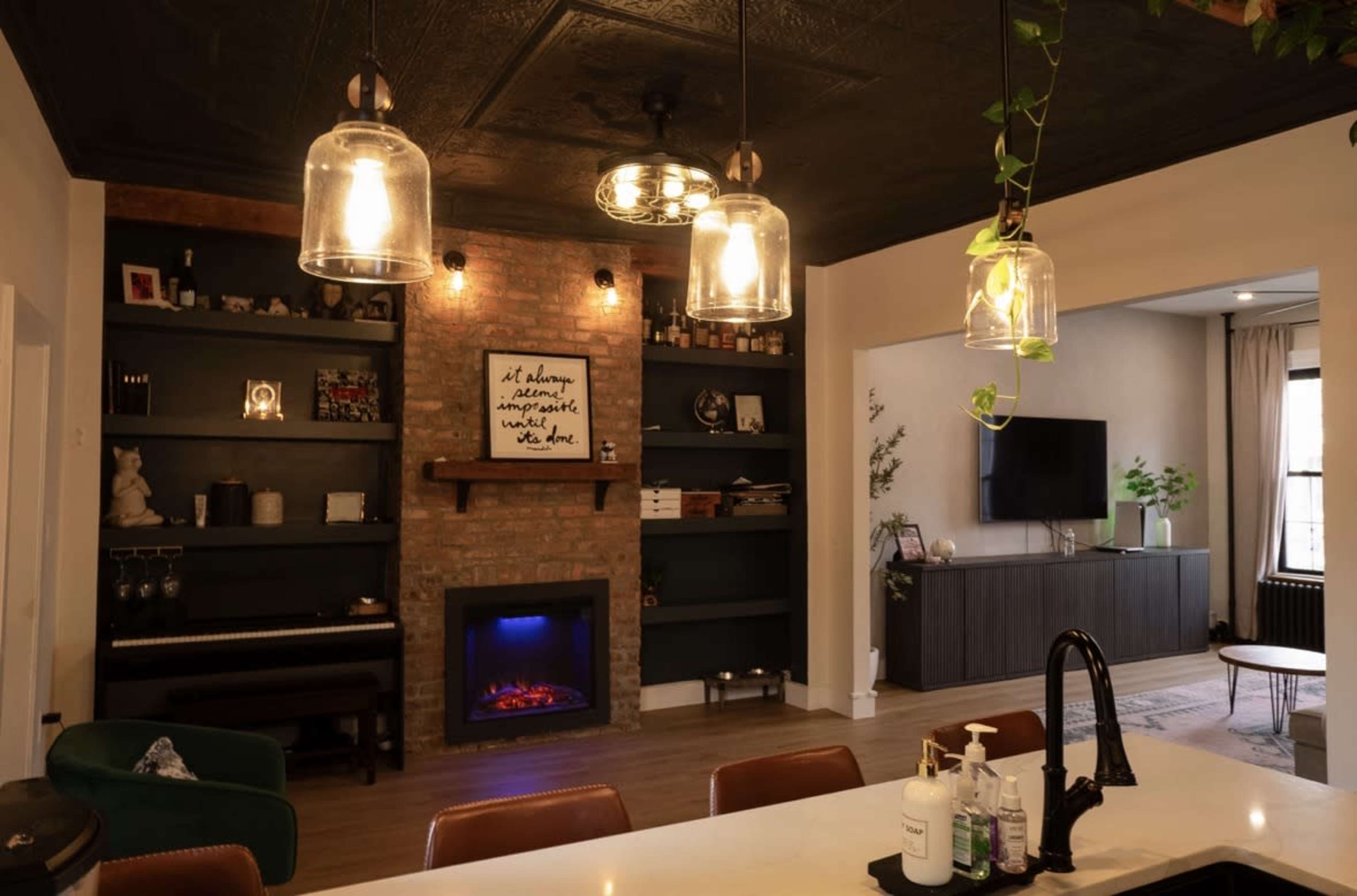 The image shows a modern living space featuring a kitchen island, an electric fireplace with a brick surround, open shelving with decorative items, and pendant lighting above the island.