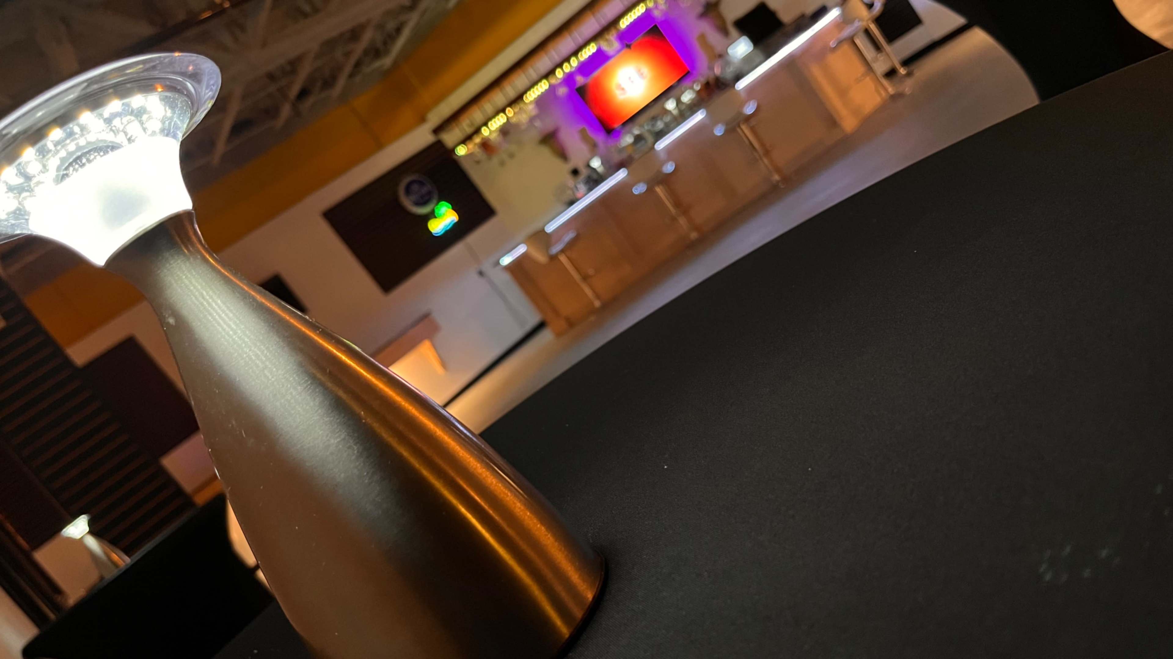A dimly lit bar area with a modern metallic lamp in the foreground and a brightly colored illuminated backdrop featuring a screen and bar counter in the background.