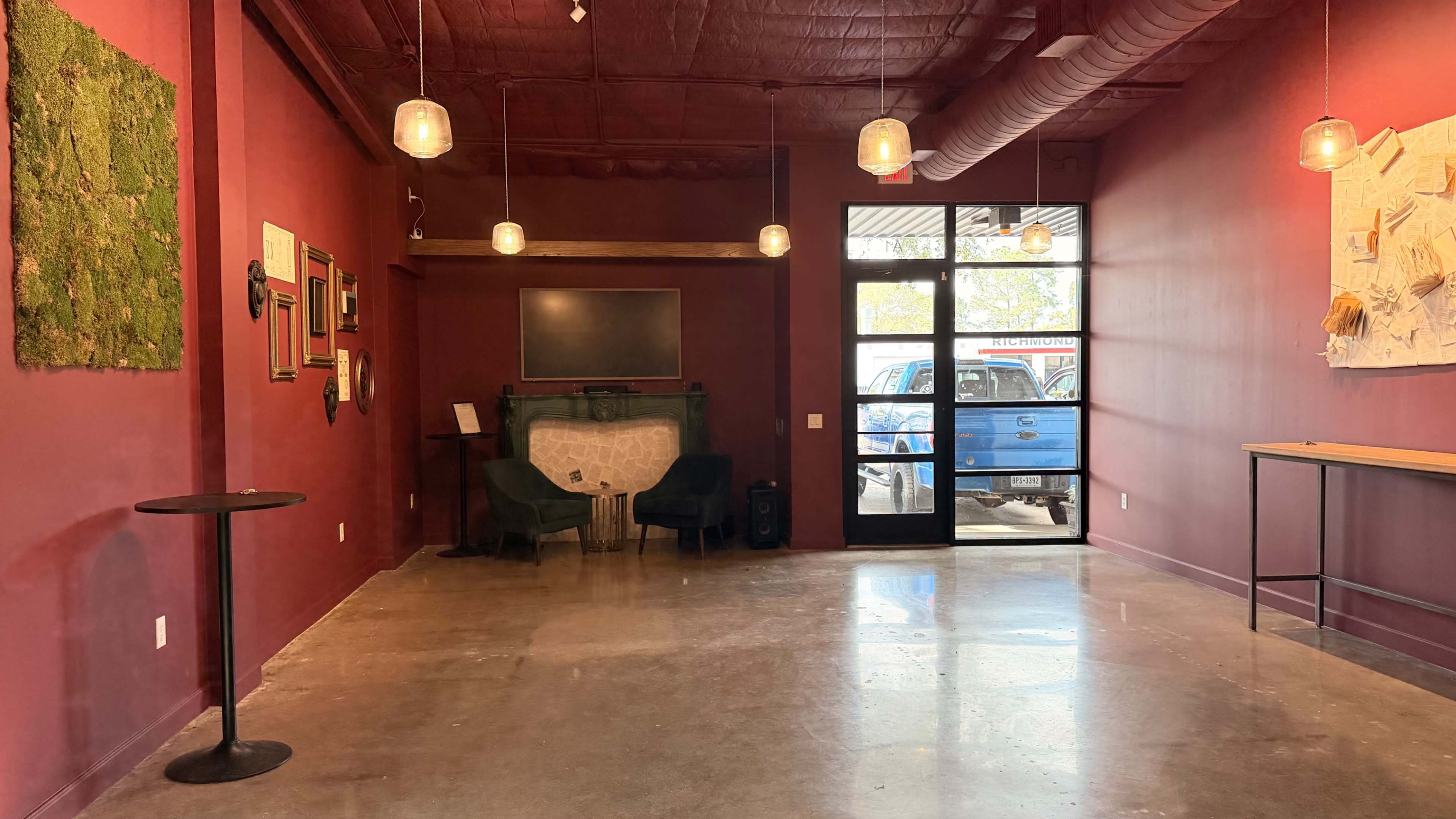 The image shows a vacant interior room with a maroon wall, two green chairs, a countertop along one side, and a large glass door facing a blue truck parked outside.