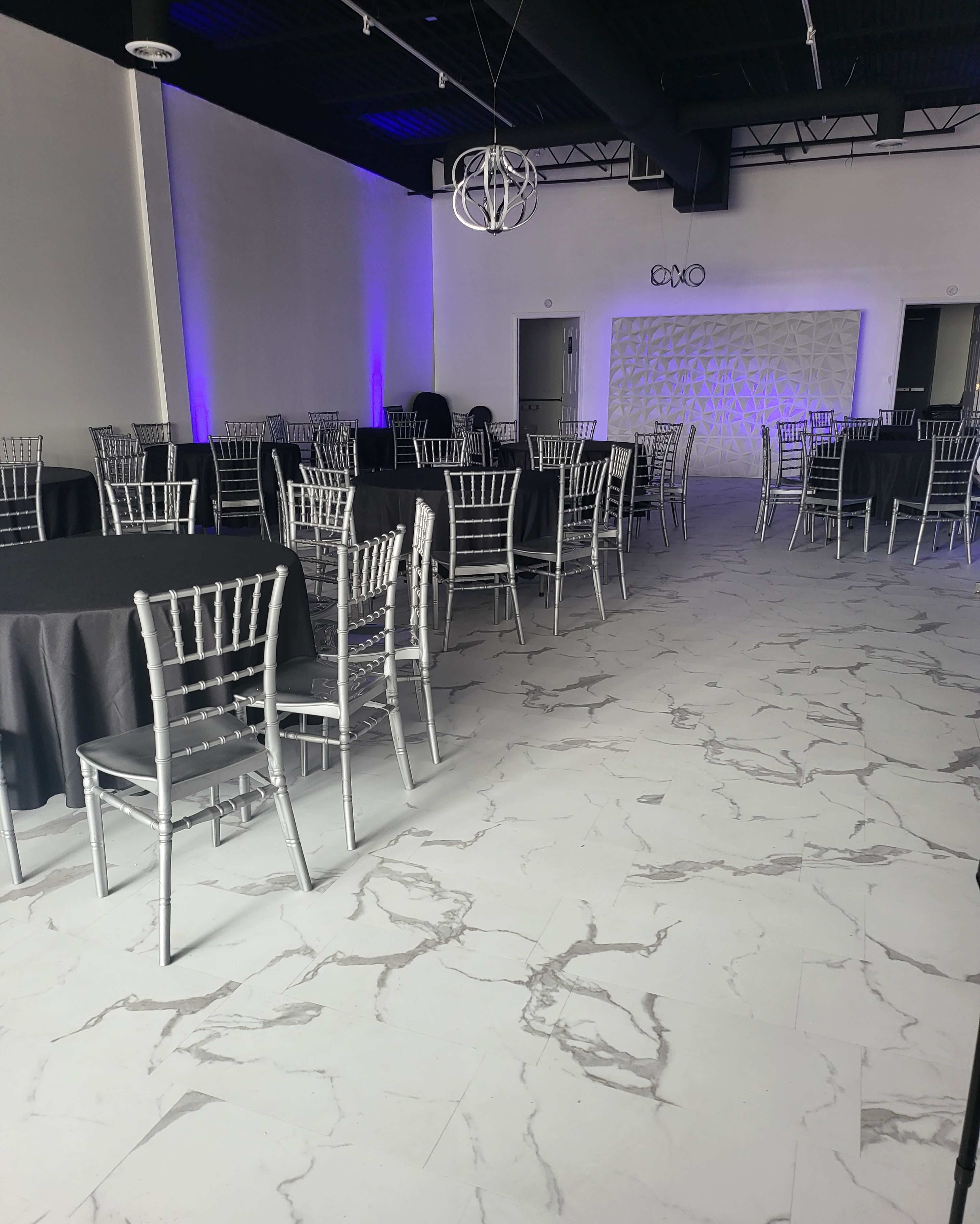 A spacious event venue with round black tables and silver chairs arranged neatly on a marble-patterned floor, illuminated by purple lighting.