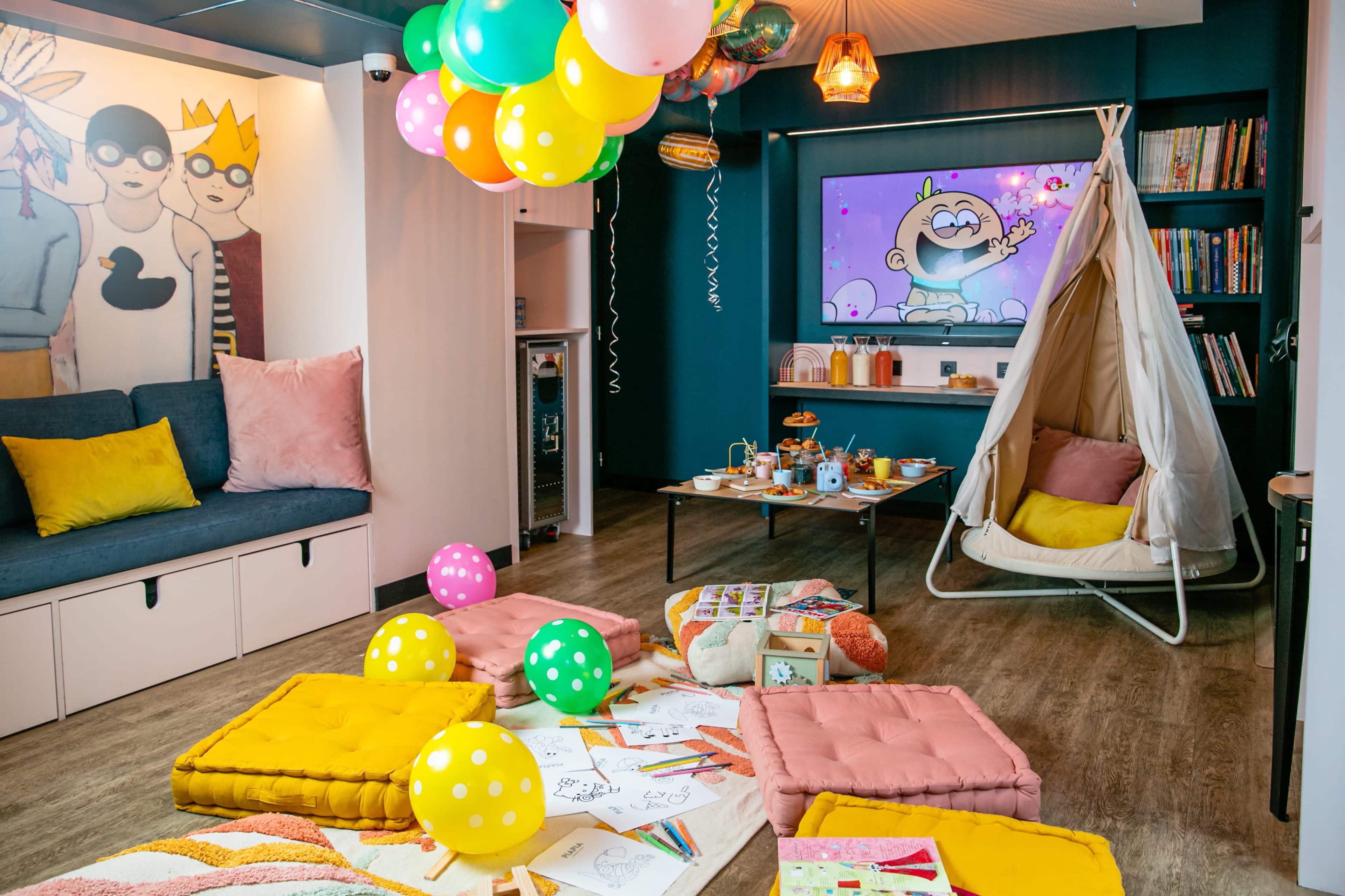 A colorful children's playroom features a swinging chair, various plush cushions on the floor, and a decorated table with snacks and art supplies, all under balloons and a television displaying animated content.