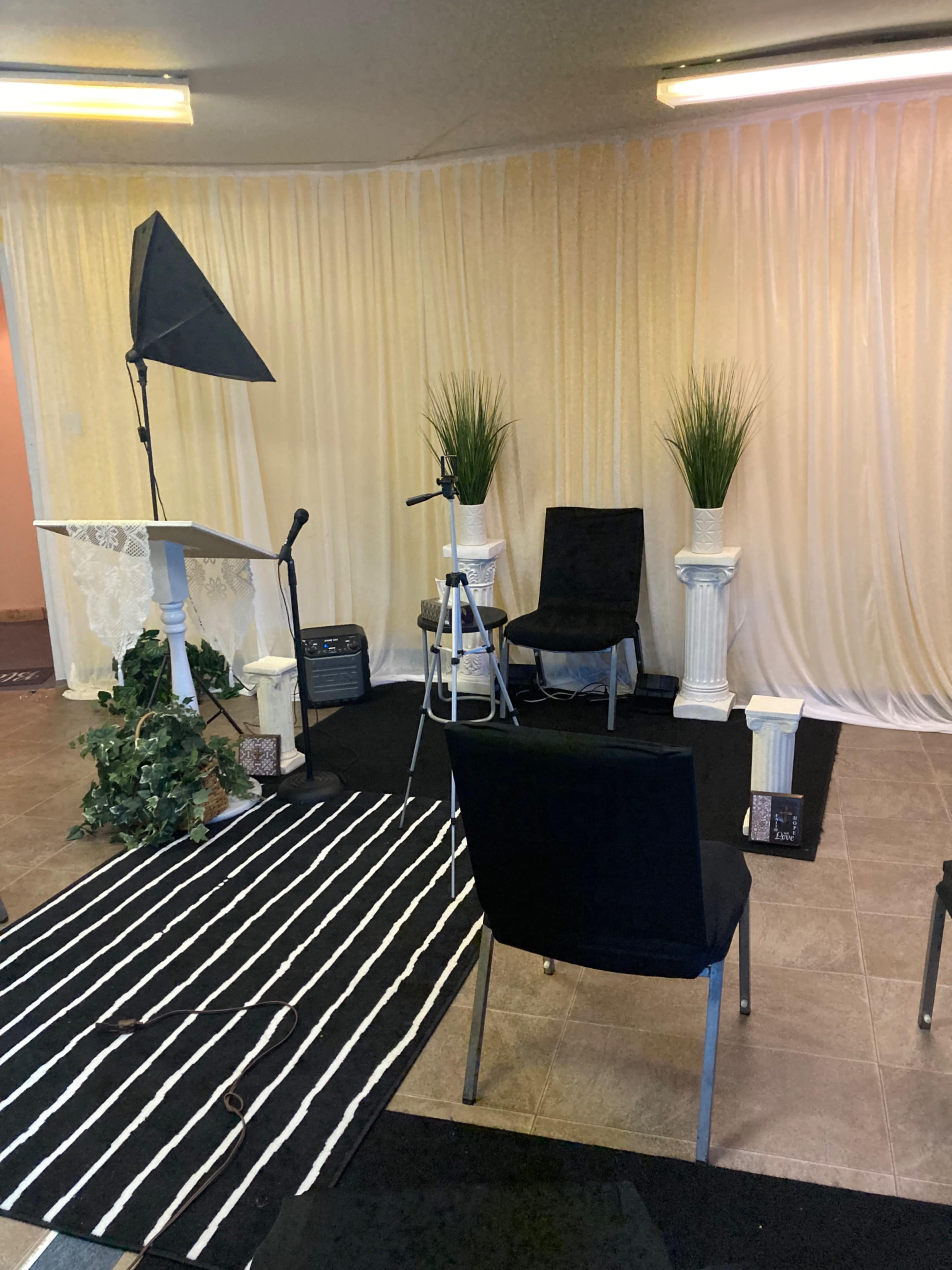 A small indoor setting arranged for a presentation or recording, featuring a podium, a microphone, a camera mounted on a tripod, two black chairs, potted plants, and a striped rug on the floor.