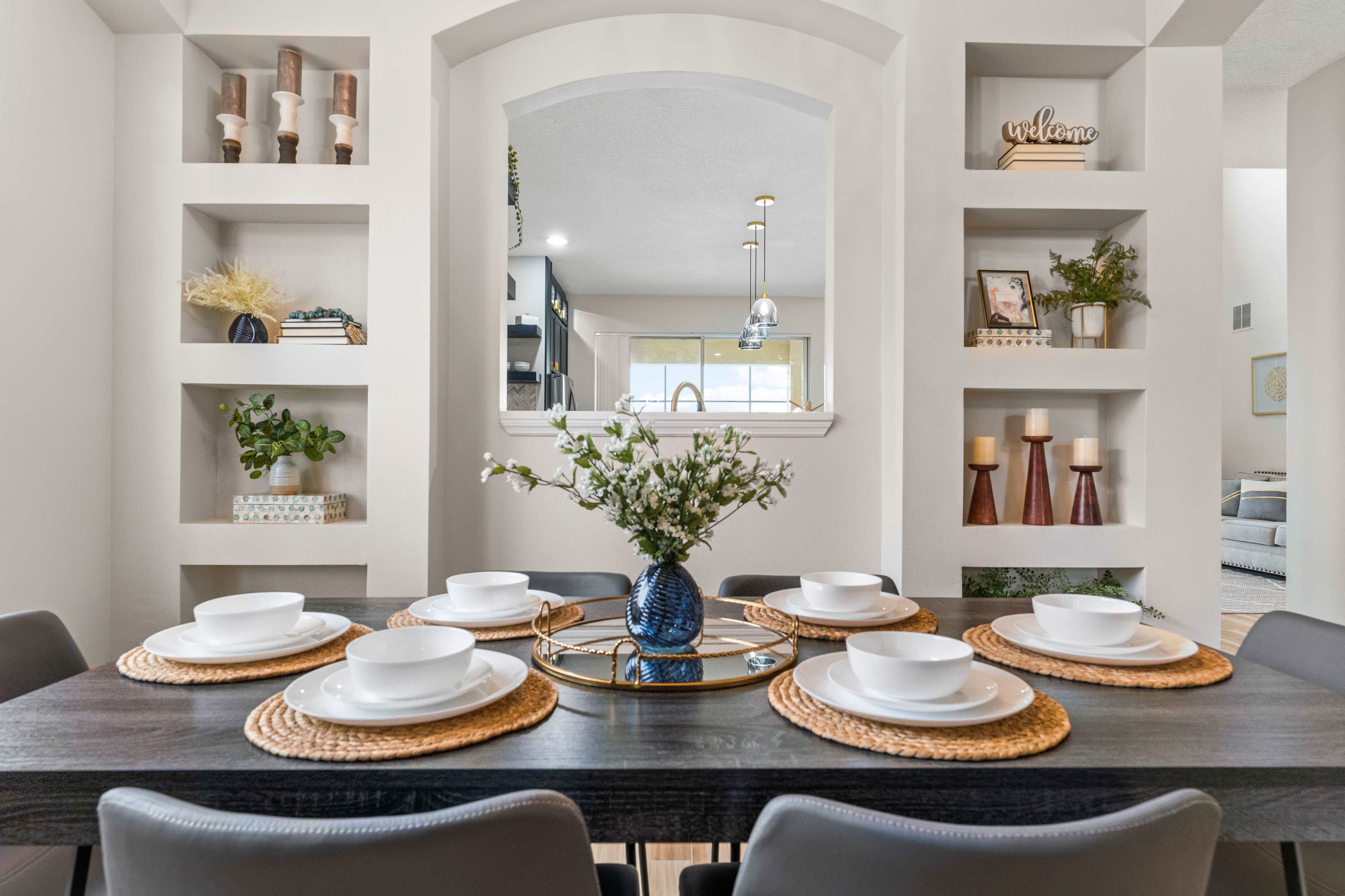 A dining table is set with six white plates on woven placemats, featuring a decorative vase of flowers at the center, surrounded by built-in shelves displaying various decorative items.