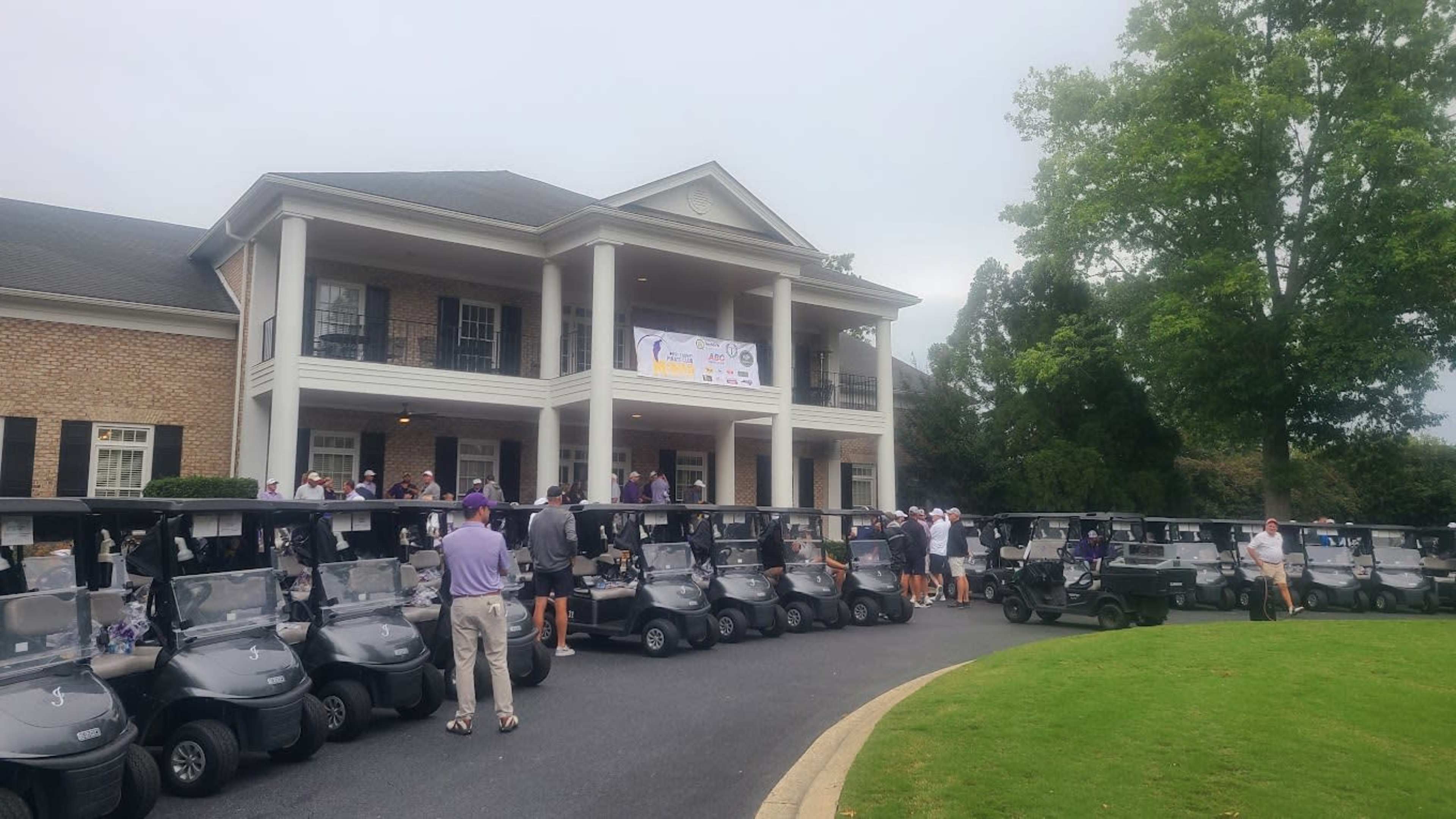 A row of golf carts is lined up in front of a brick building with a porch, where people are gathered for an event.