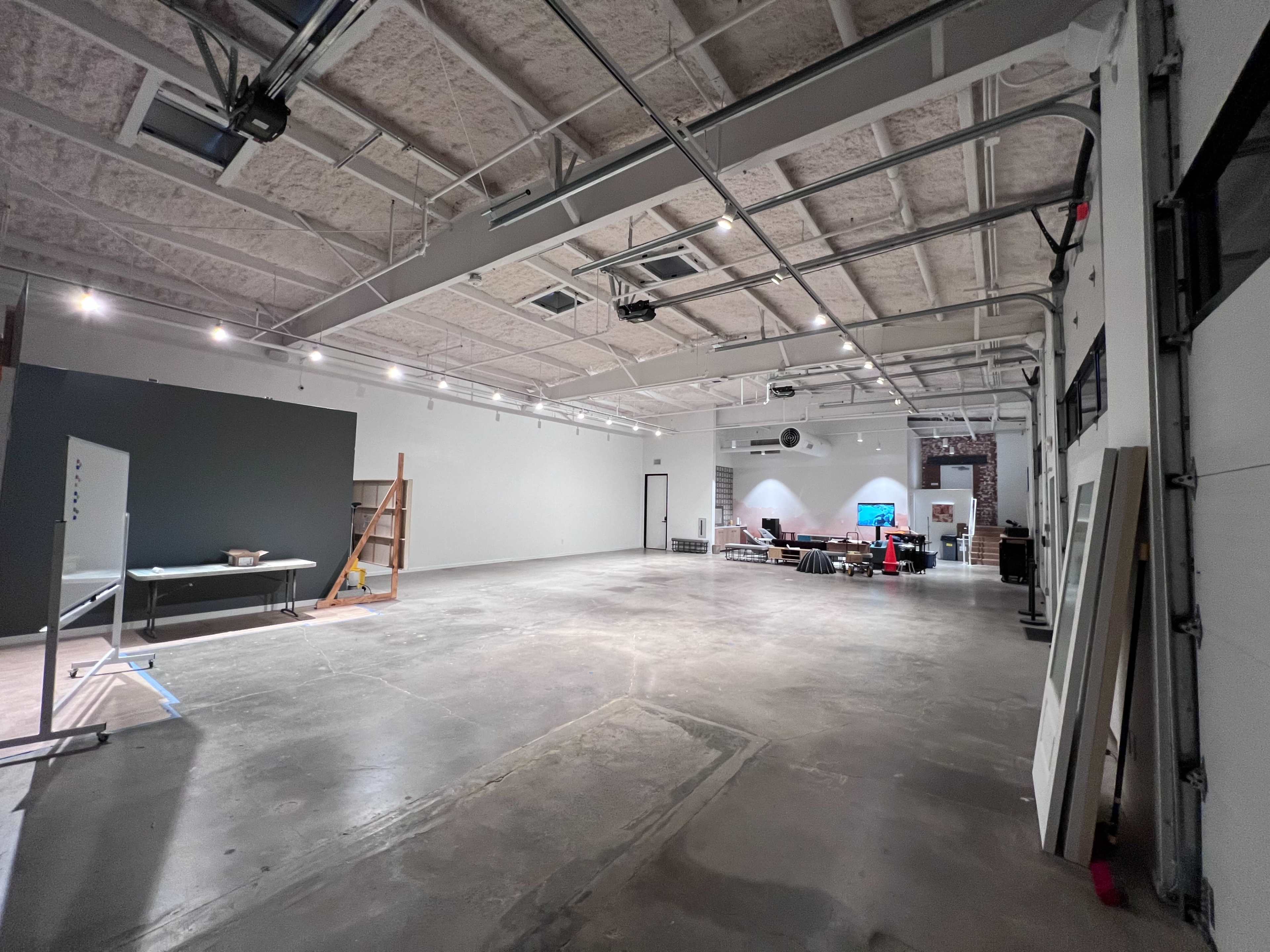 The image shows a spacious, empty industrial room with high ceilings, concrete flooring, and various furniture and equipment in the background.