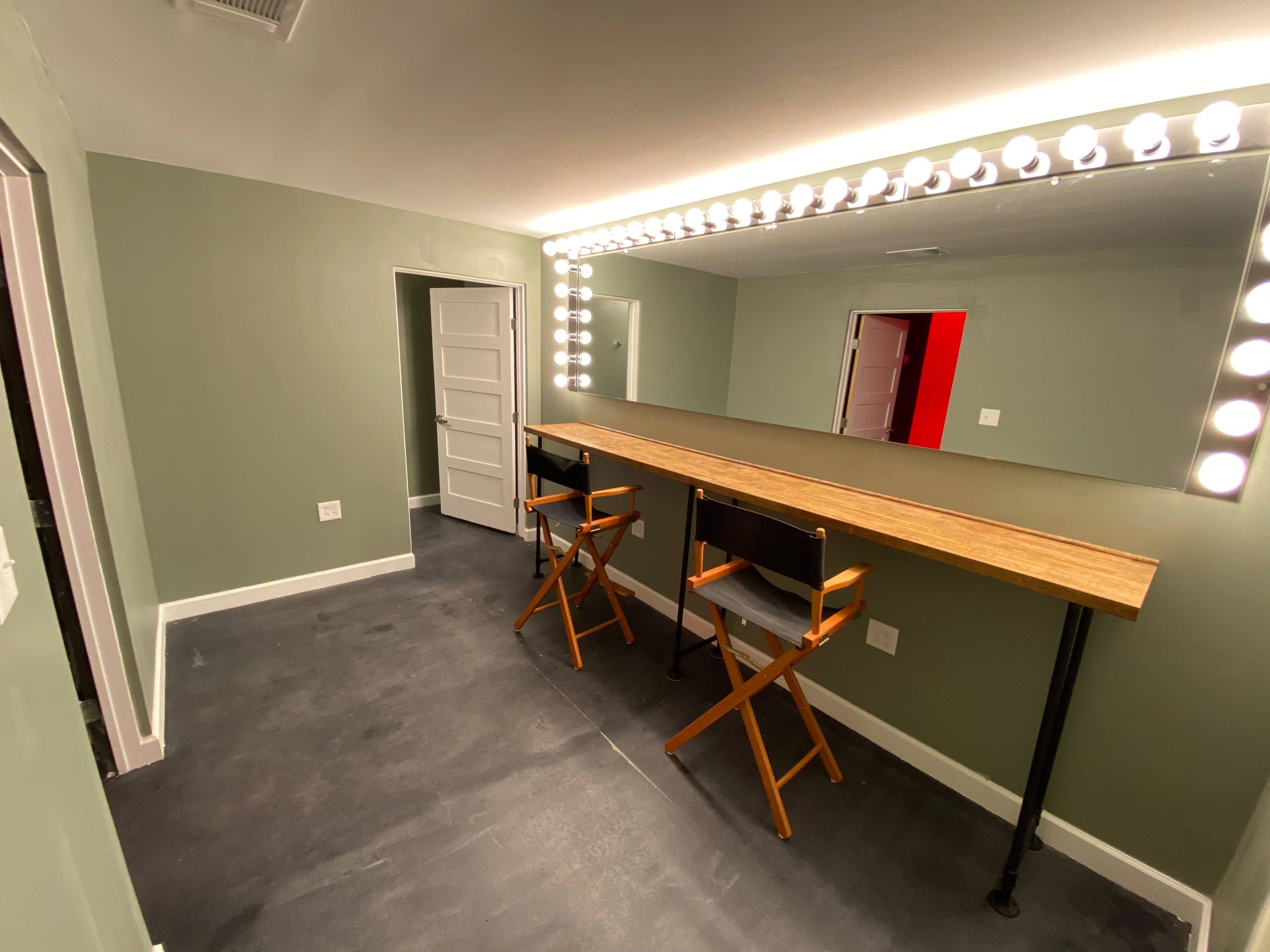A room with a green-painted wall, a long wooden table with two director's chairs, and a large mirror illuminated by bright, round light bulbs.