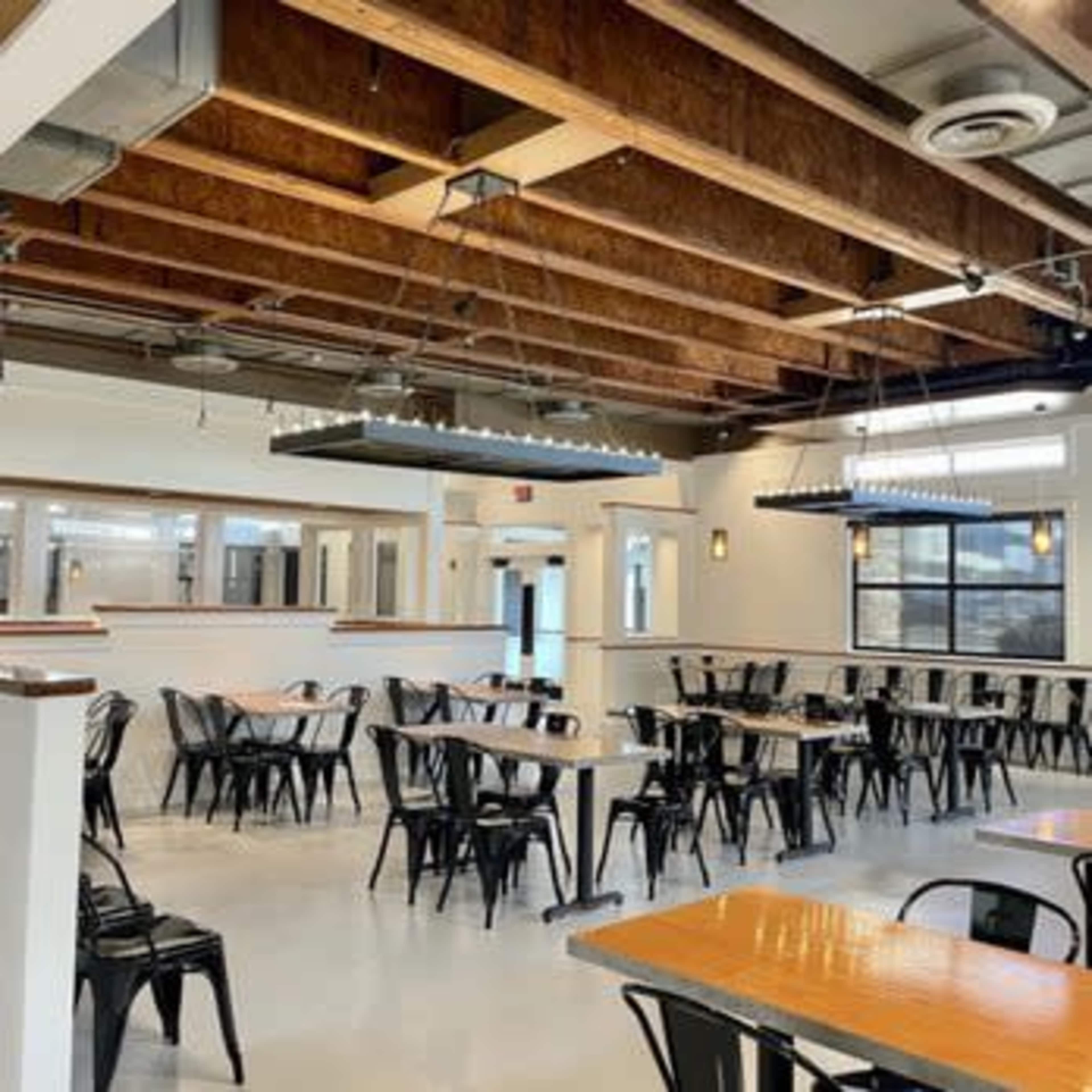 The image shows a spacious dining area with metal chairs and wooden tables, featuring exposed beams and modern lighting fixtures.