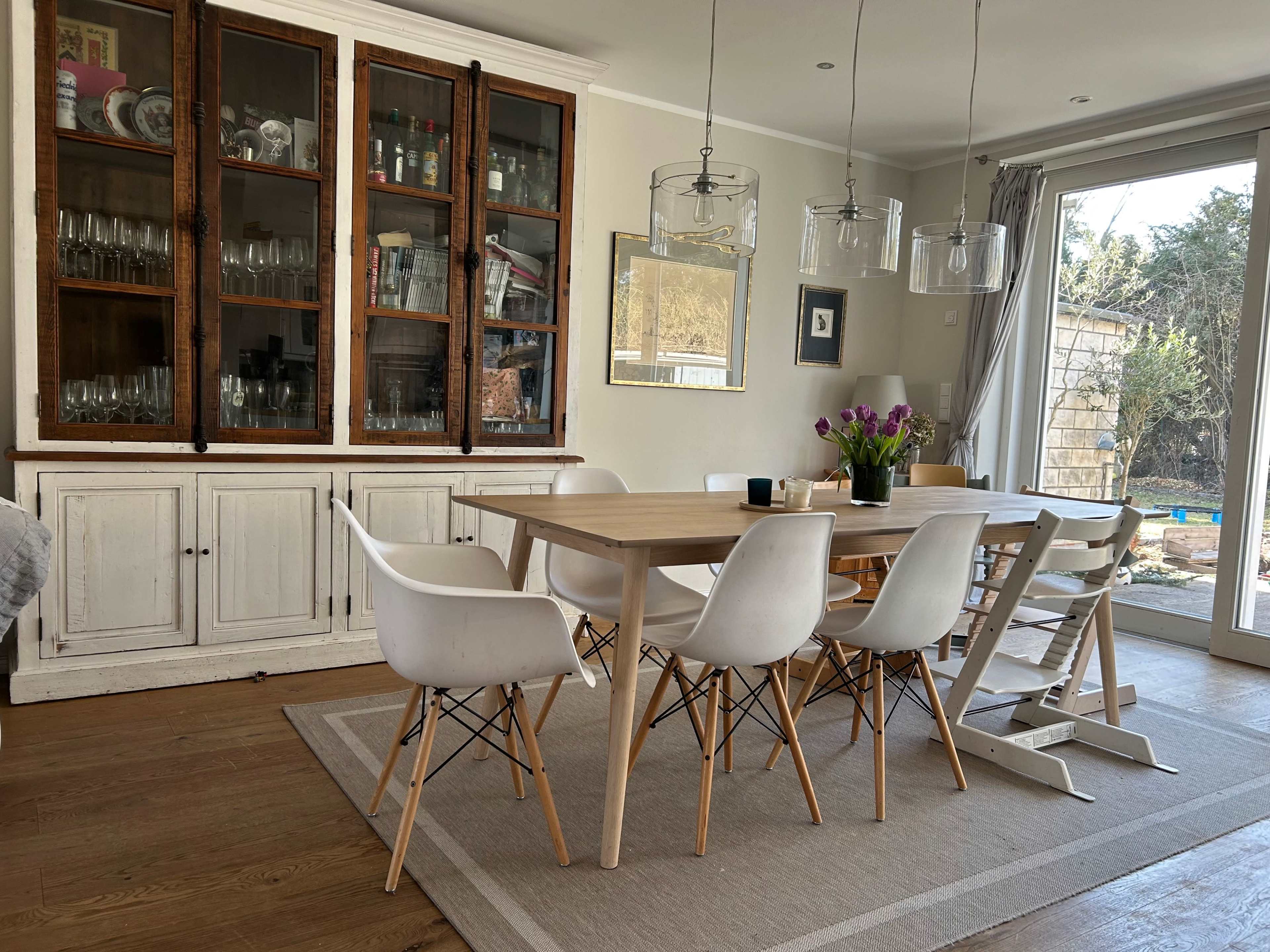 A dining room features a large wooden table surrounded by white chairs, with a glass-front cabinet displaying kitchenware and natural light coming through a sliding door.