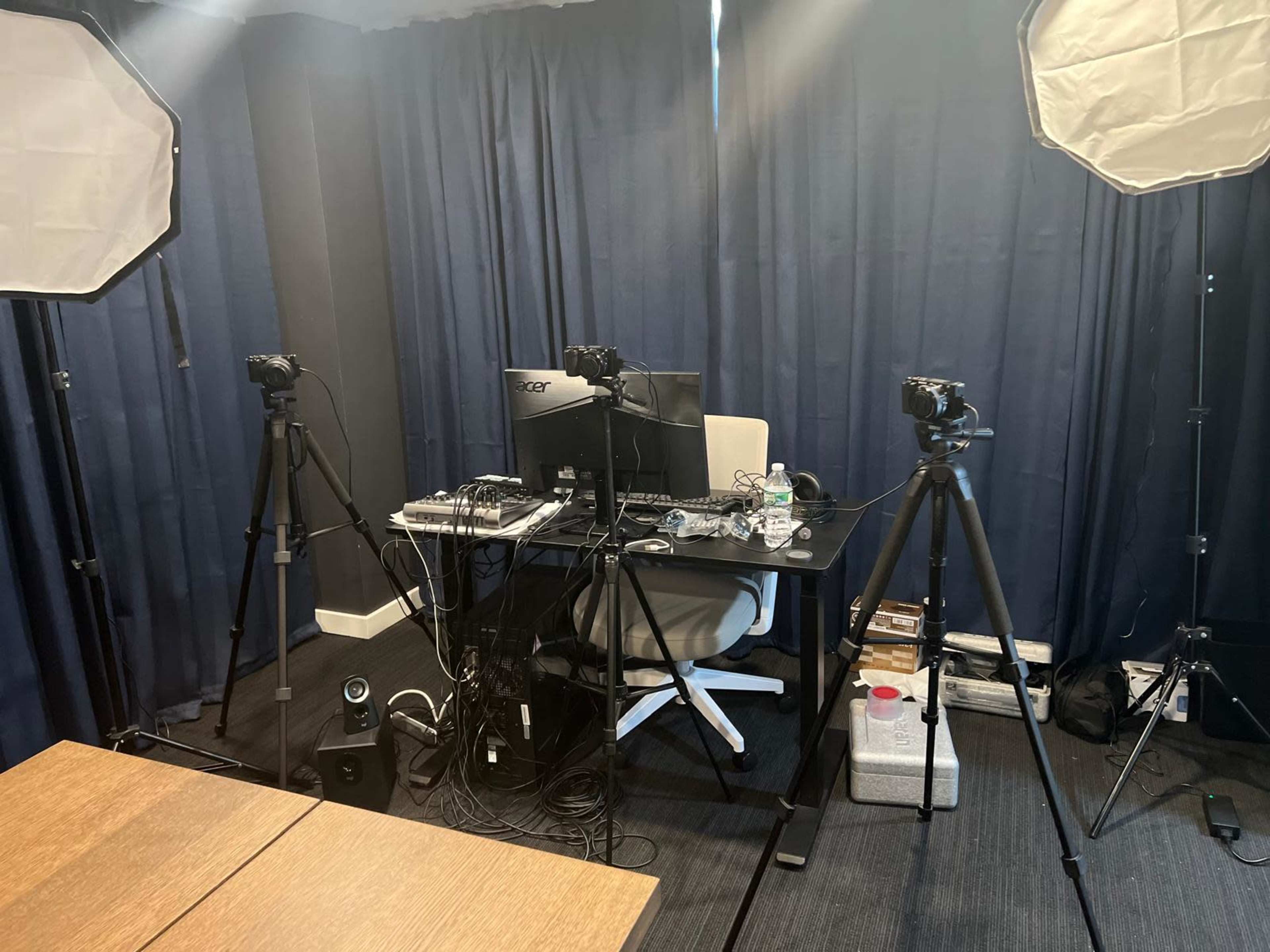 The image shows a video recording setup with a desk, computer, and multiple cameras on tripods, surrounded by softbox lights and blue curtains.