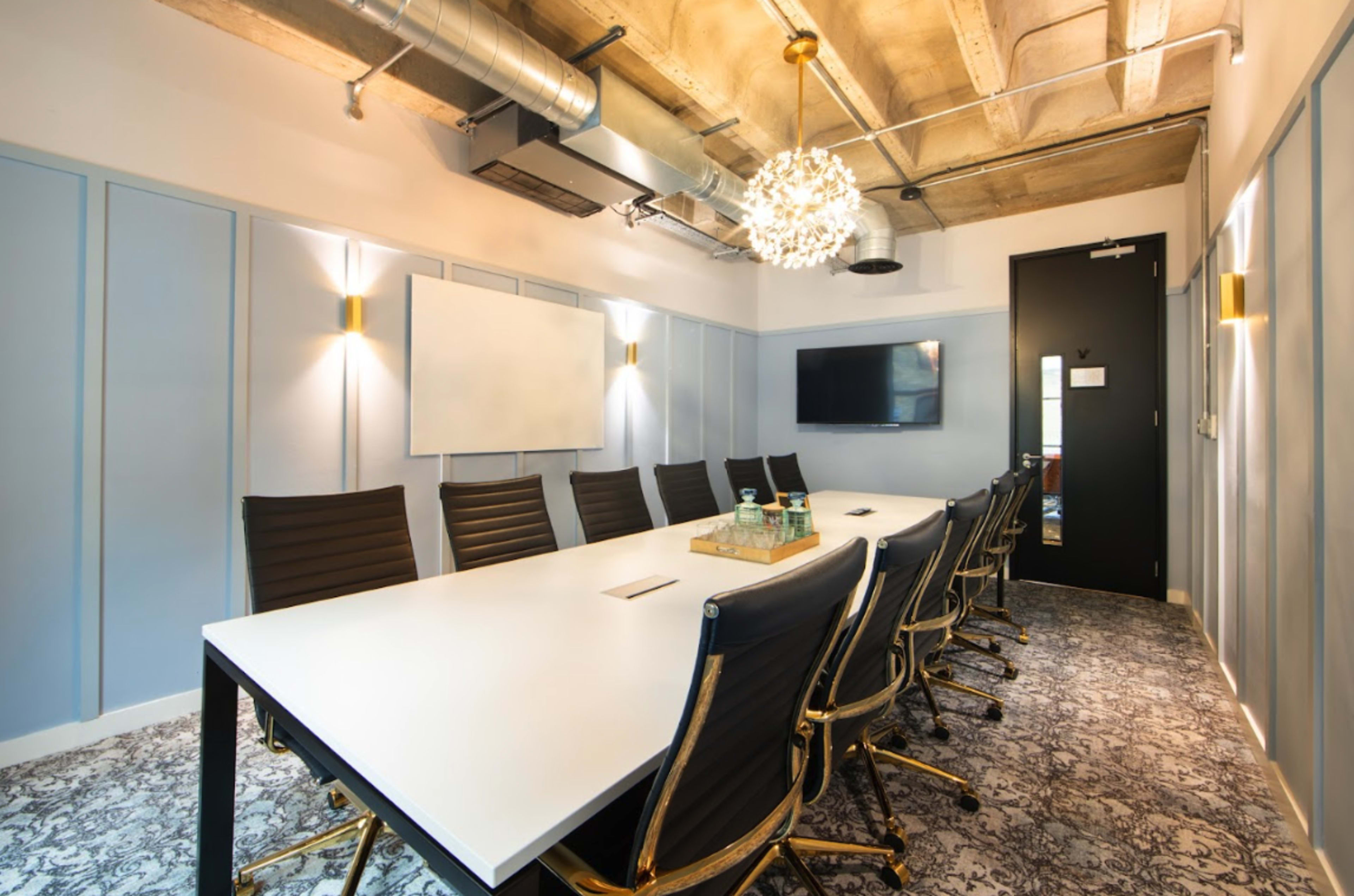A modern conference room featuring a long rectangular table surrounded by black leather chairs, with a whiteboard and a television mounted on the wall.