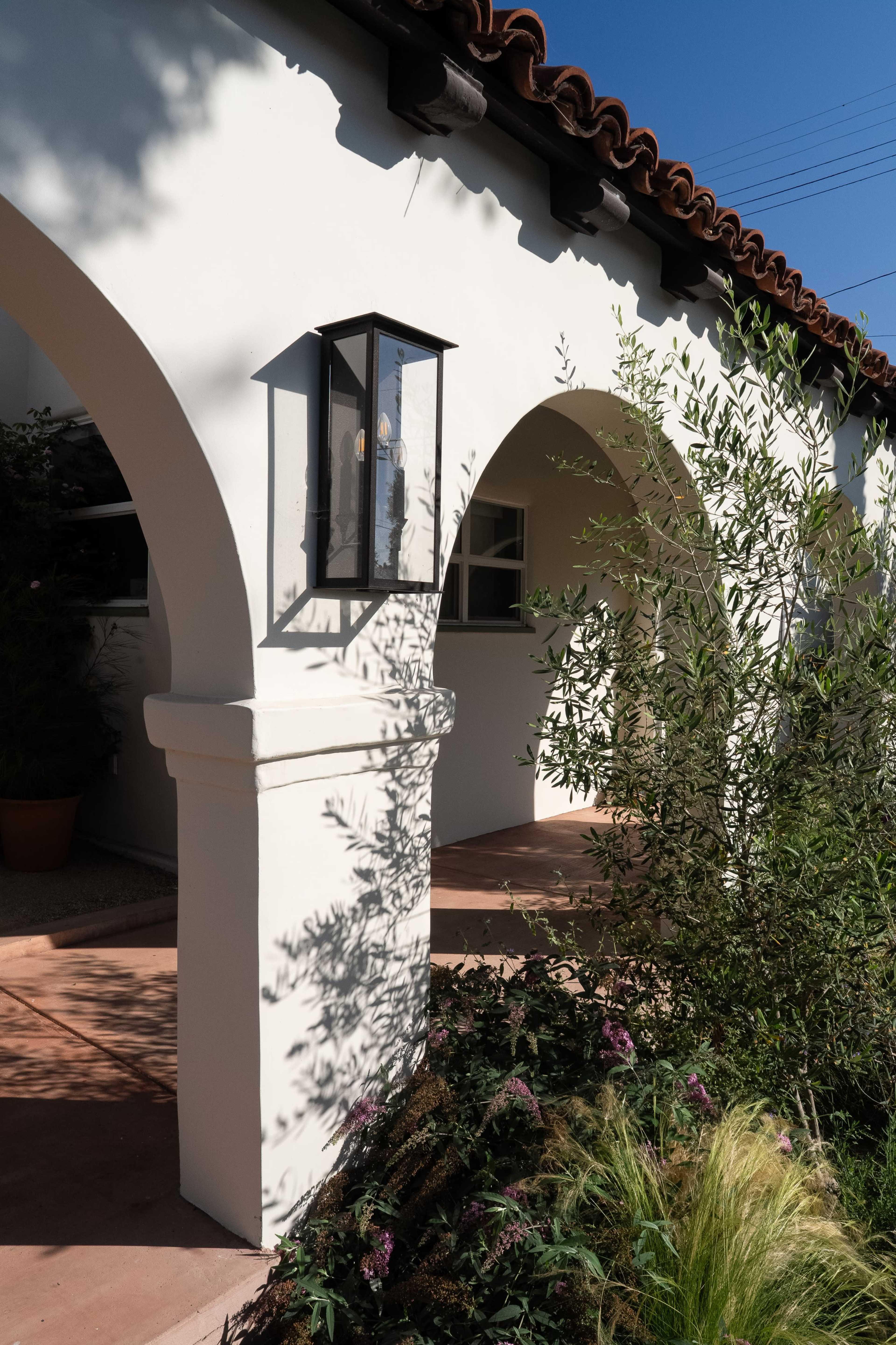 The image shows a white stucco building with arched doorways and a lantern, surrounded by greenery and colorful plants.