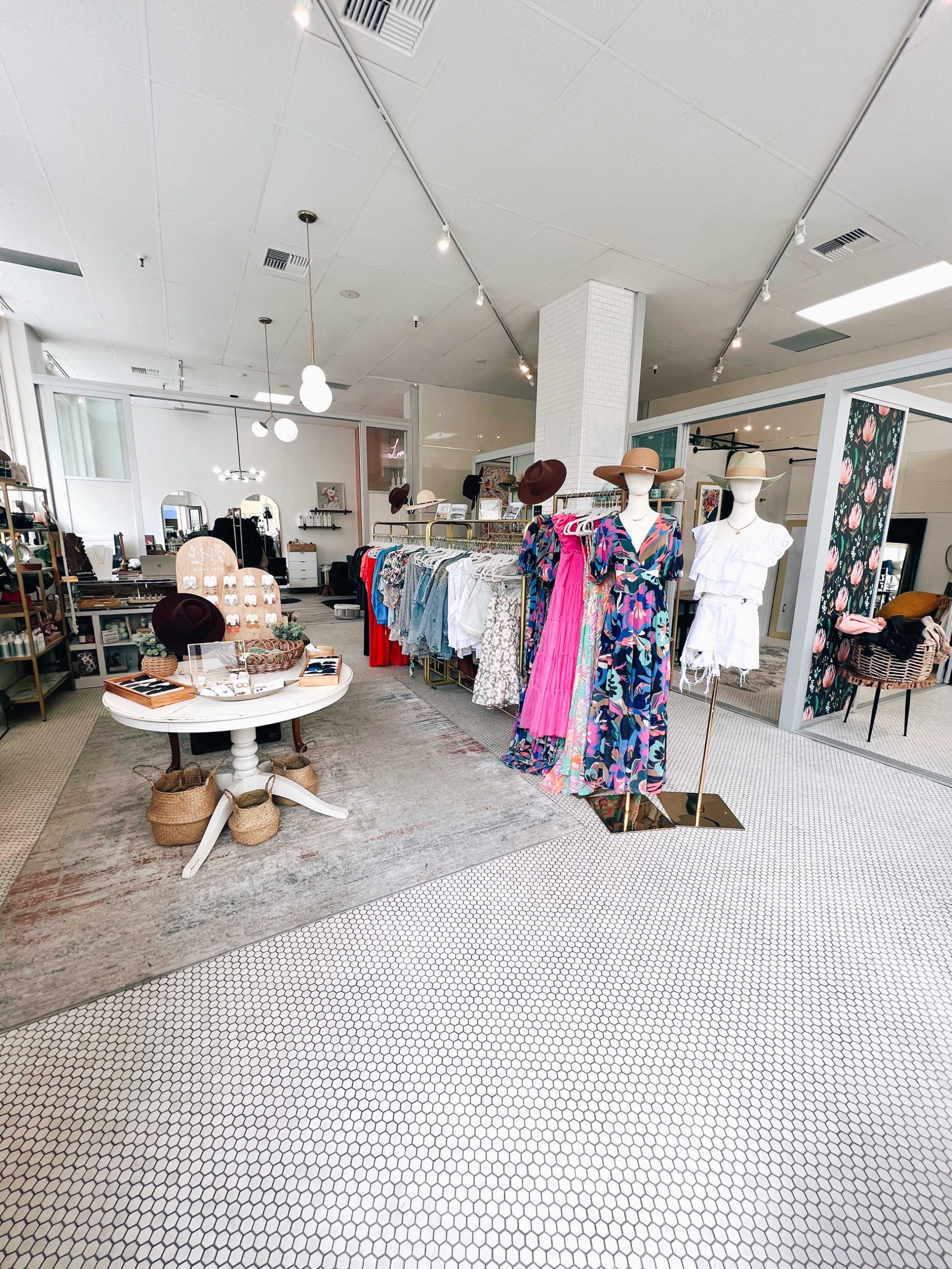 The interior of a boutique featuring clothing racks, a round table, and decorative elements, with a light-colored tile floor and bright lighting.