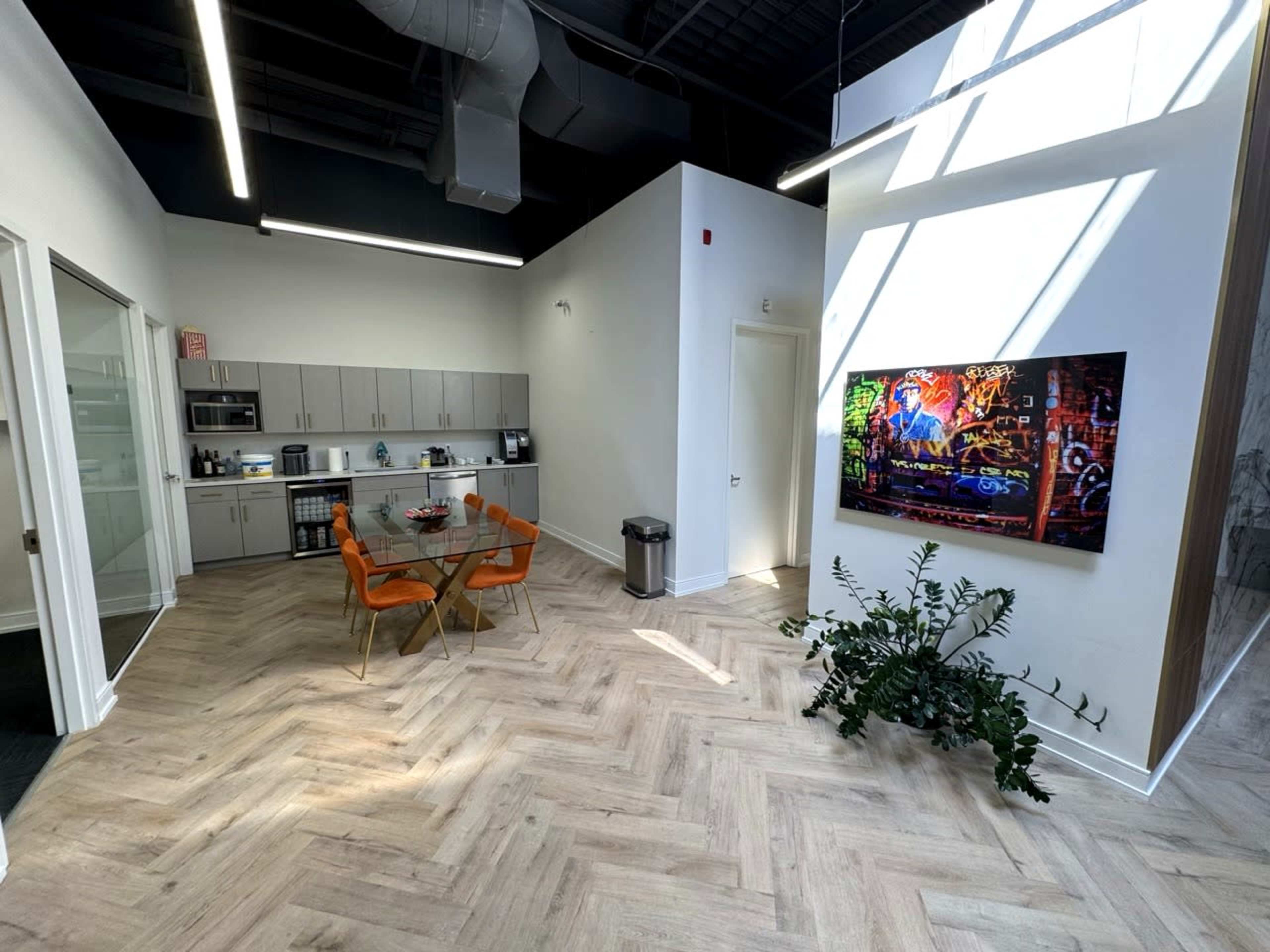 The image shows a modern office interior featuring a kitchenette, a circular table with orange chairs, and a TV mounted on the wall, with natural light streaming in through the windows.