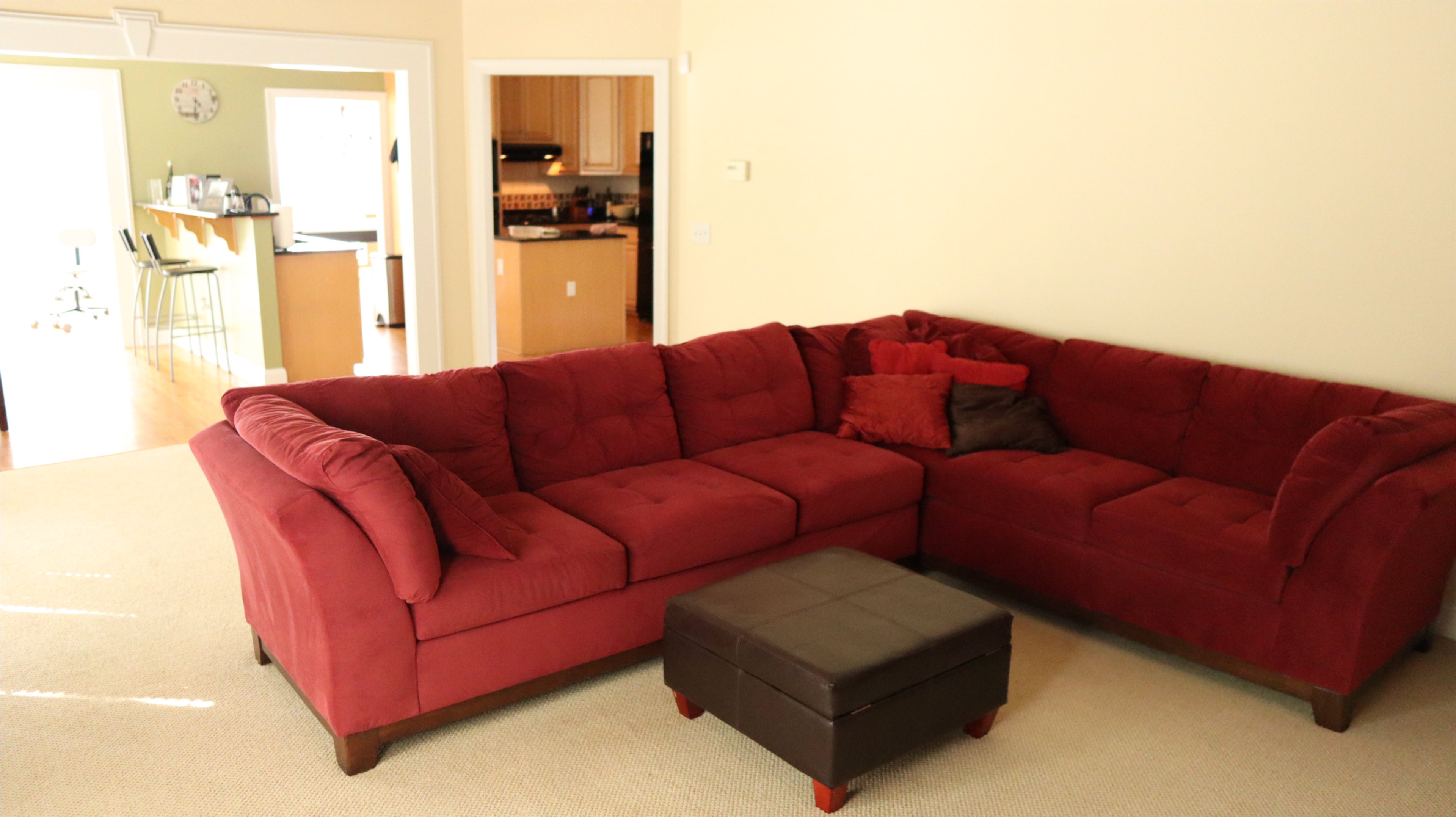 A red sectional sofa and a dark ottoman are positioned in a light-filled living room adjacent to a kitchen area.