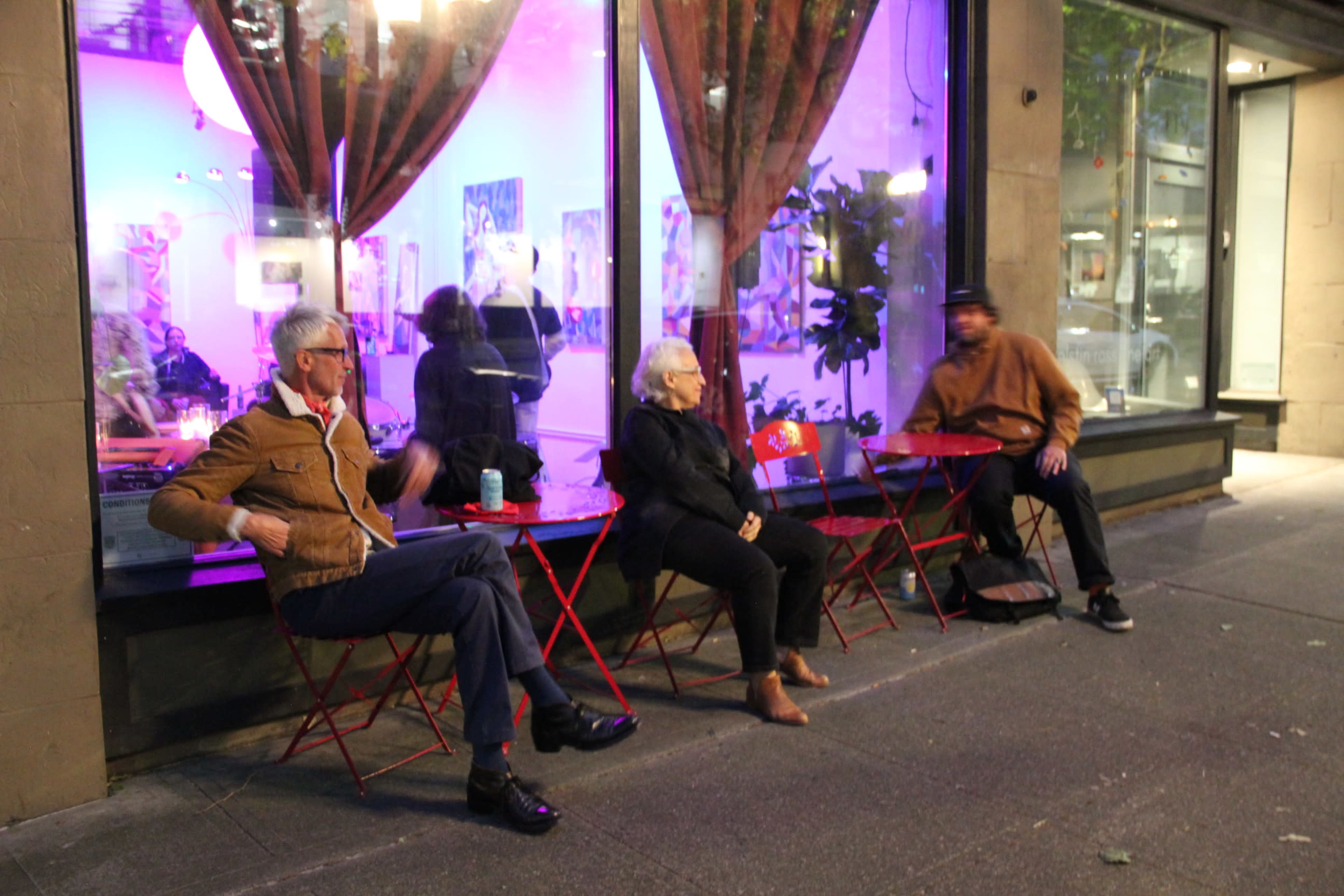Three individuals sit on red foldable chairs outside a storefront with purple lighting and curtains.