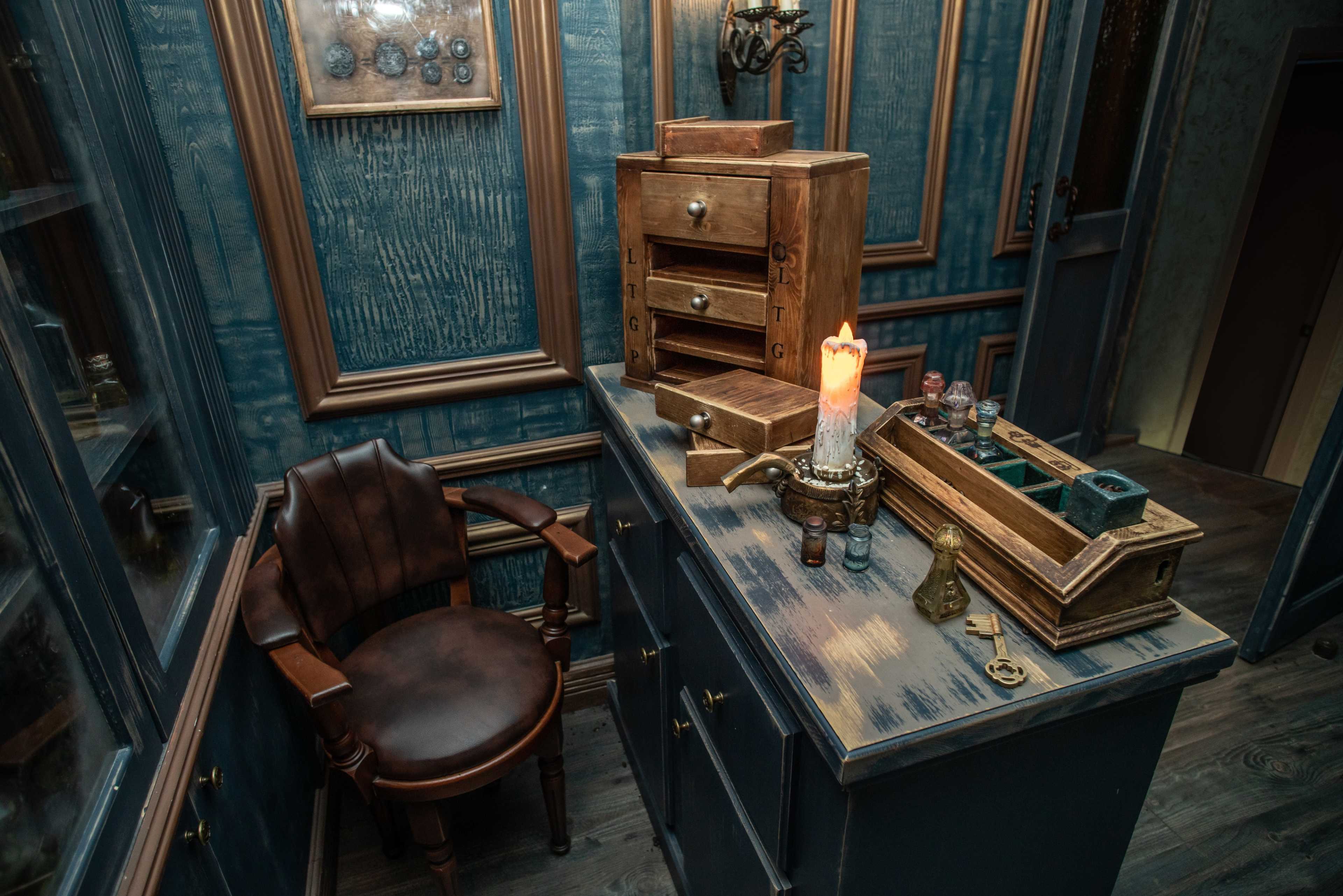 A wooden desk with various storage boxes and a lit candle sits beside a dark leather chair in a dimly lit room.