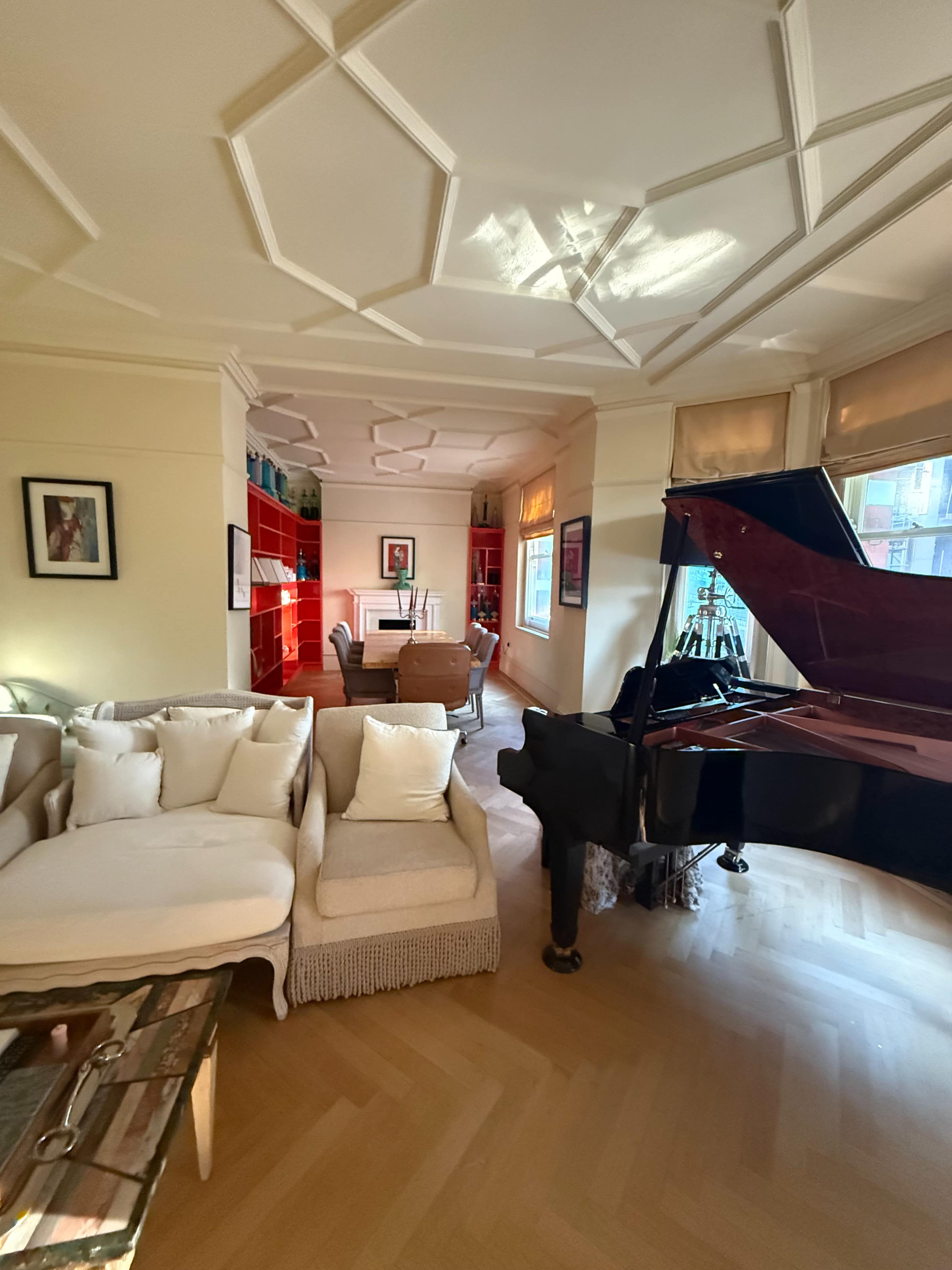 The image shows a well-lit living room featuring a grand piano, a light-colored sofa, and red shelving units along the walls.