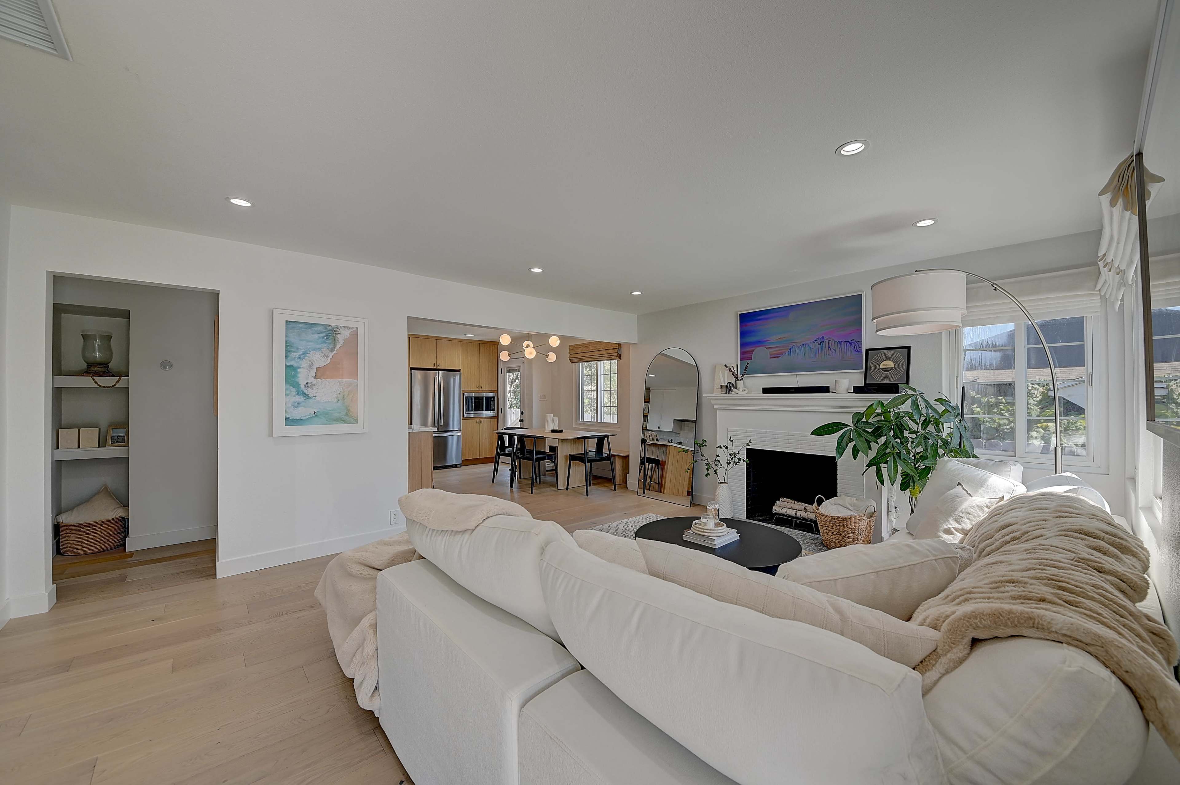 A modern, bright living room features a white sectional sofa, a coffee table, and a large window, with a dining area visible in the background.