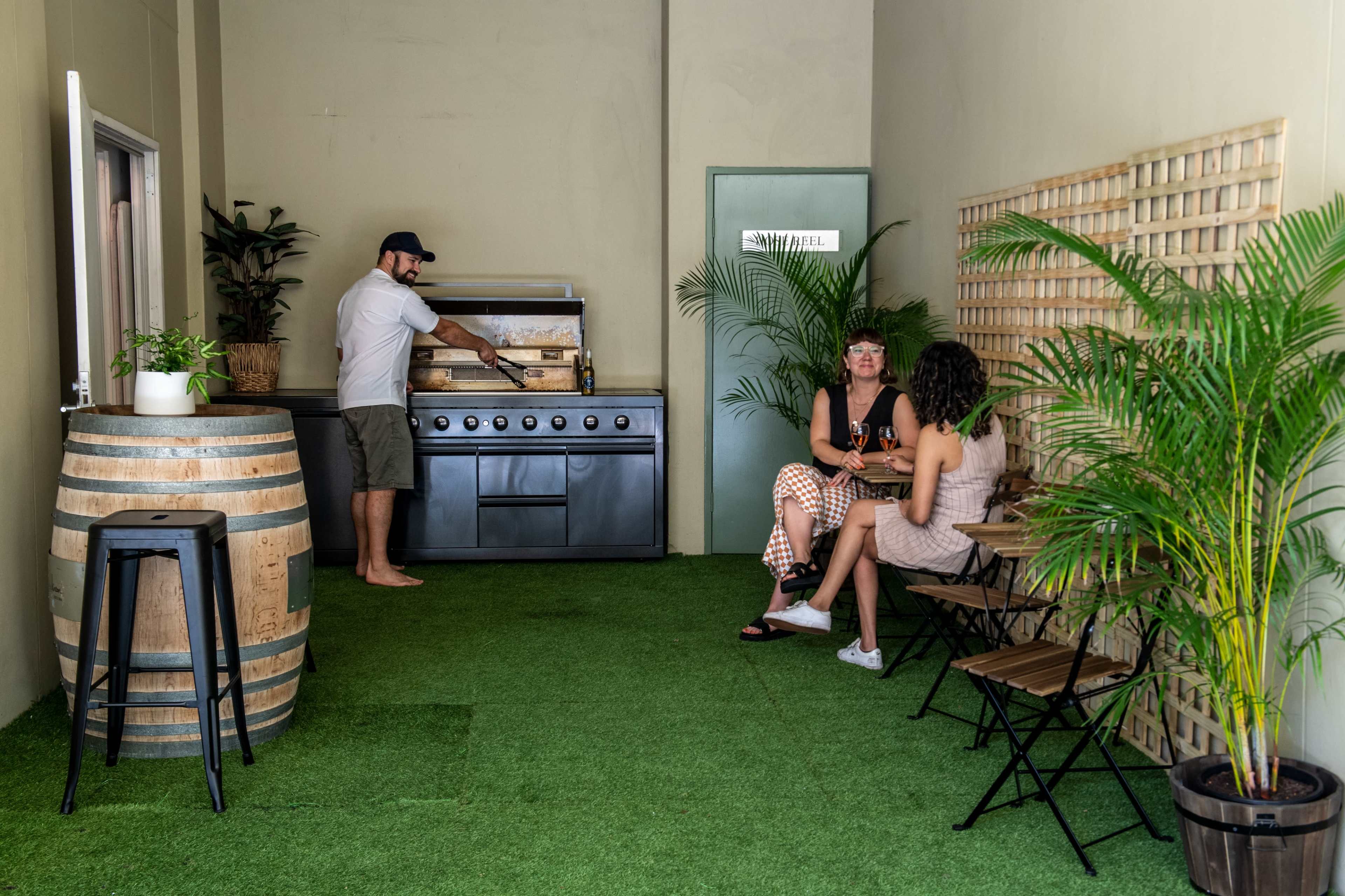 A person cooks at a grill while two women sit at a small table surrounded by potted plants in a well-lit indoor space.