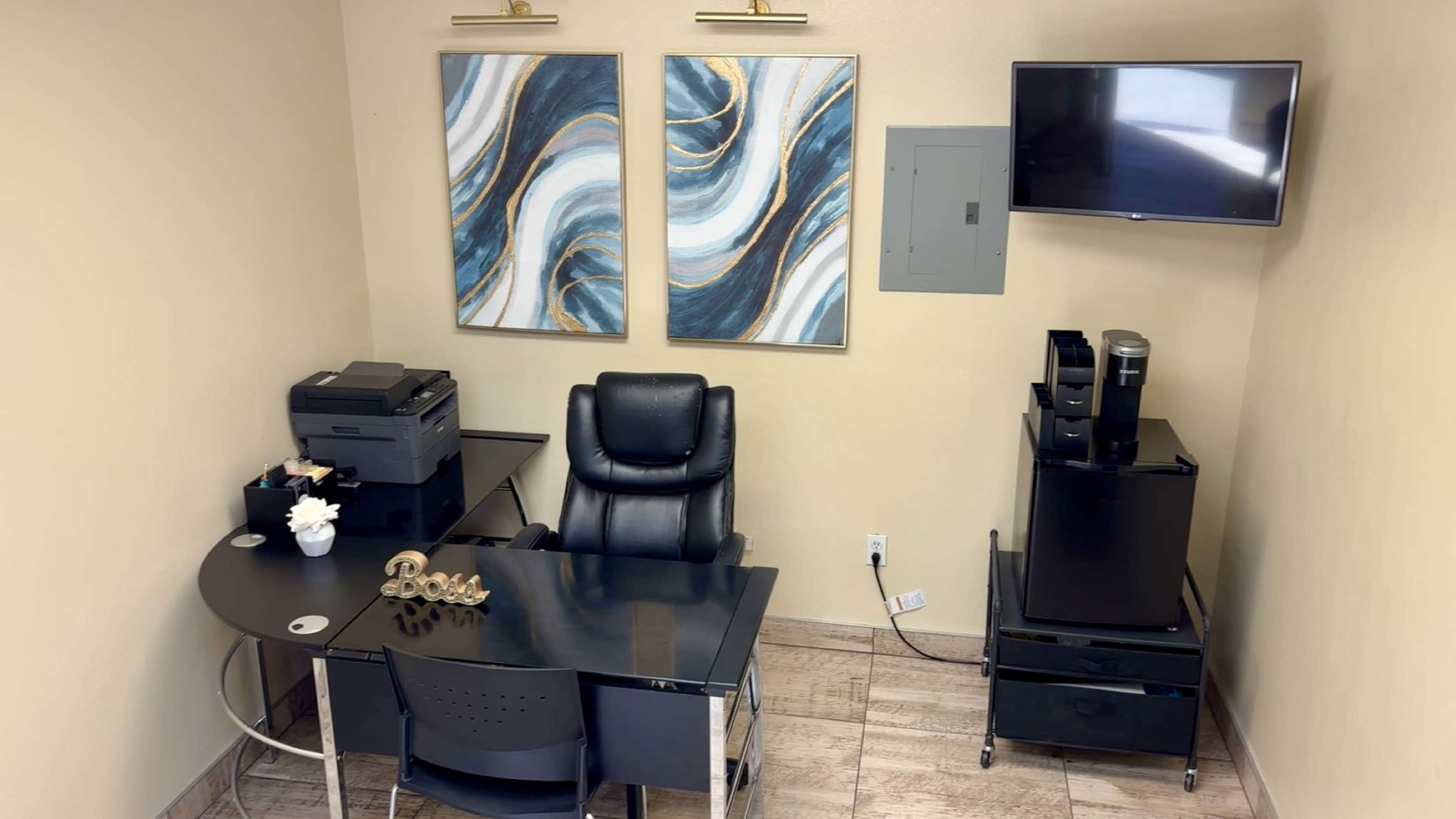 The image shows a small office space featuring a black desk with a swivel chair, a printer, decorative wall art, a mounted television, and a small mobile cart.
