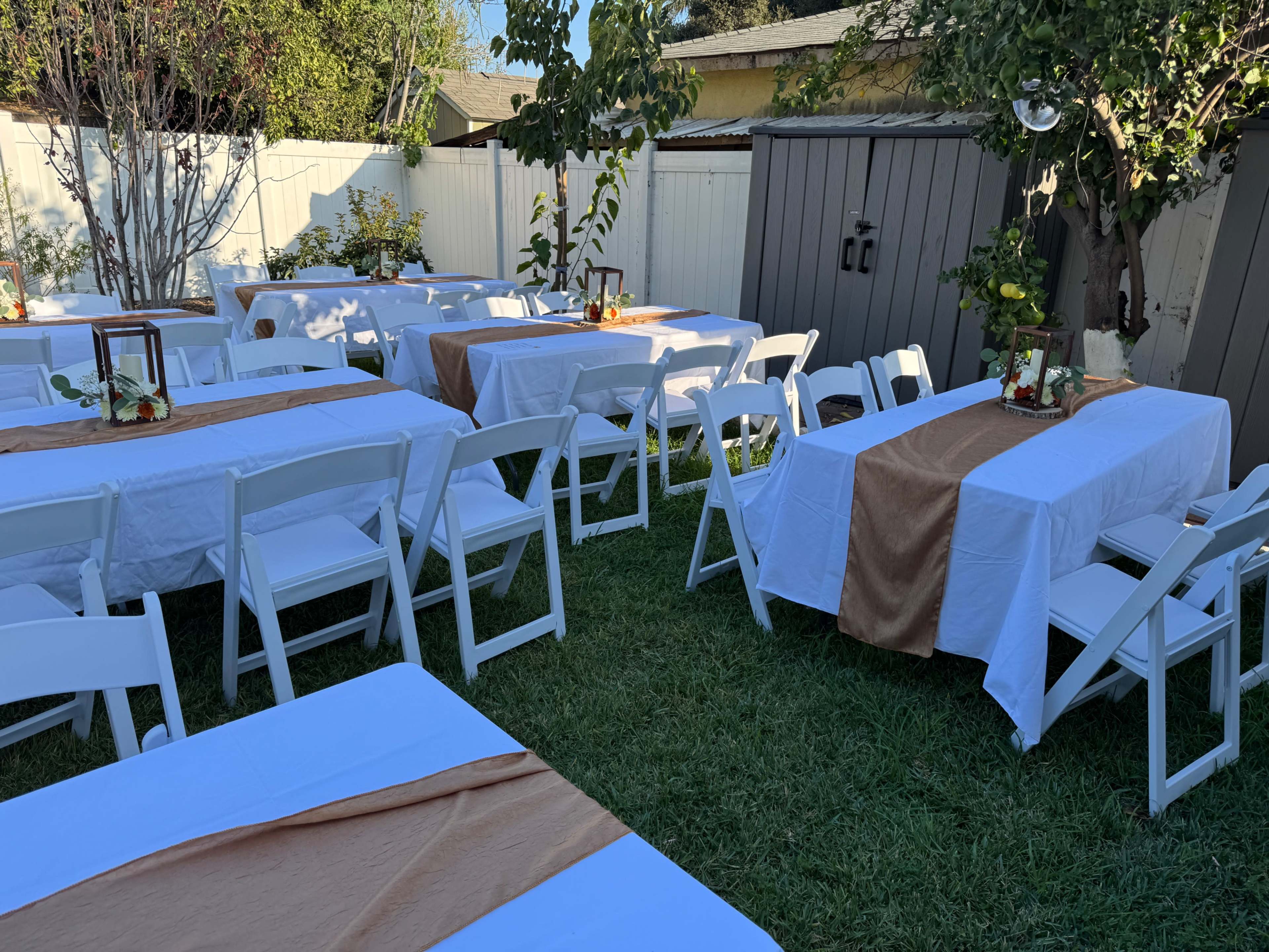 The scene features a grassy backyard set up with several white tables covered in white tablecloths, each adorned with a brown runner and centerpieces, surrounded by white folding chairs.