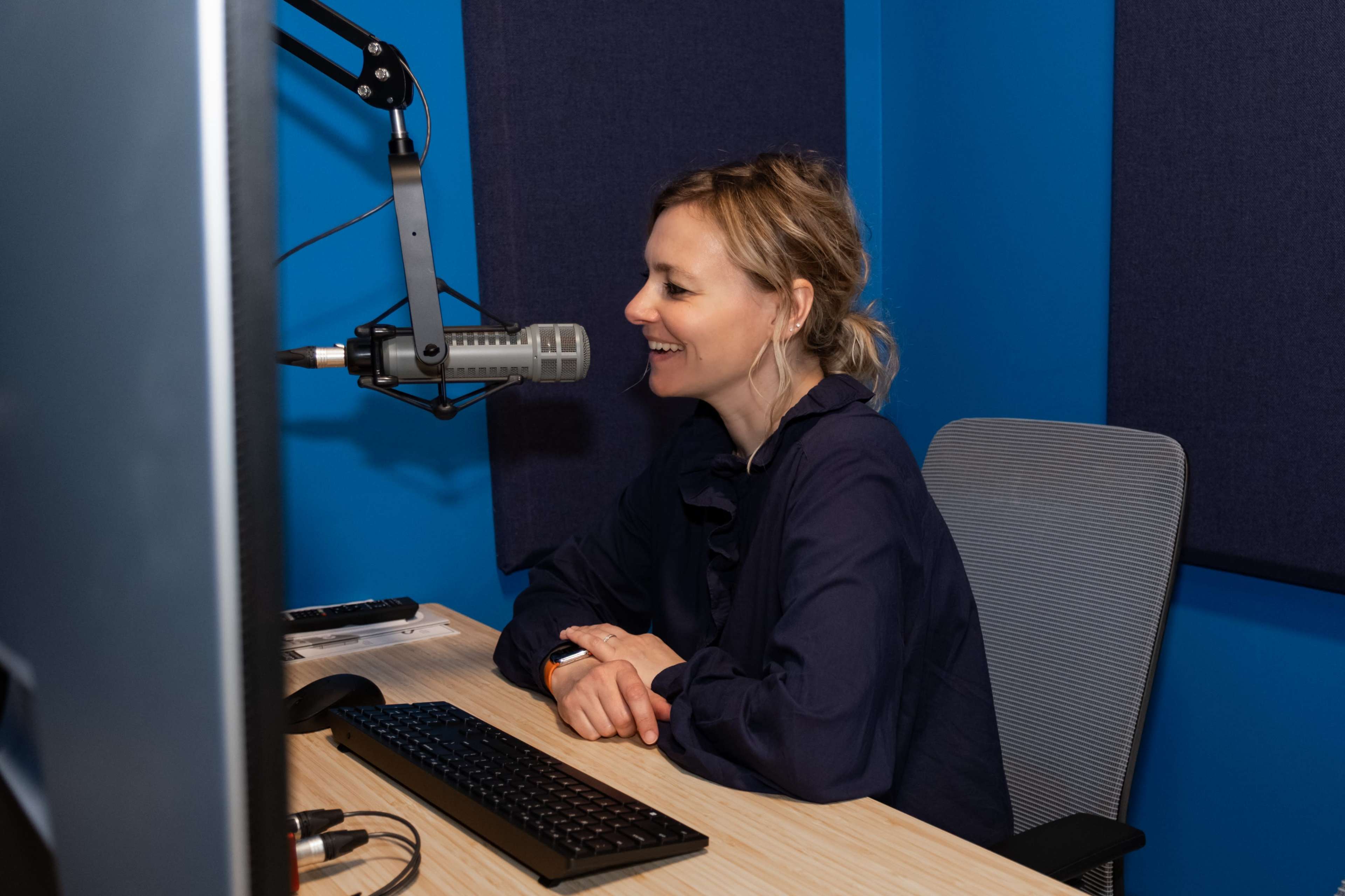 A woman sits at a desk with a computer, smiling while speaking into a microphone in a blue-walled recording studio.
