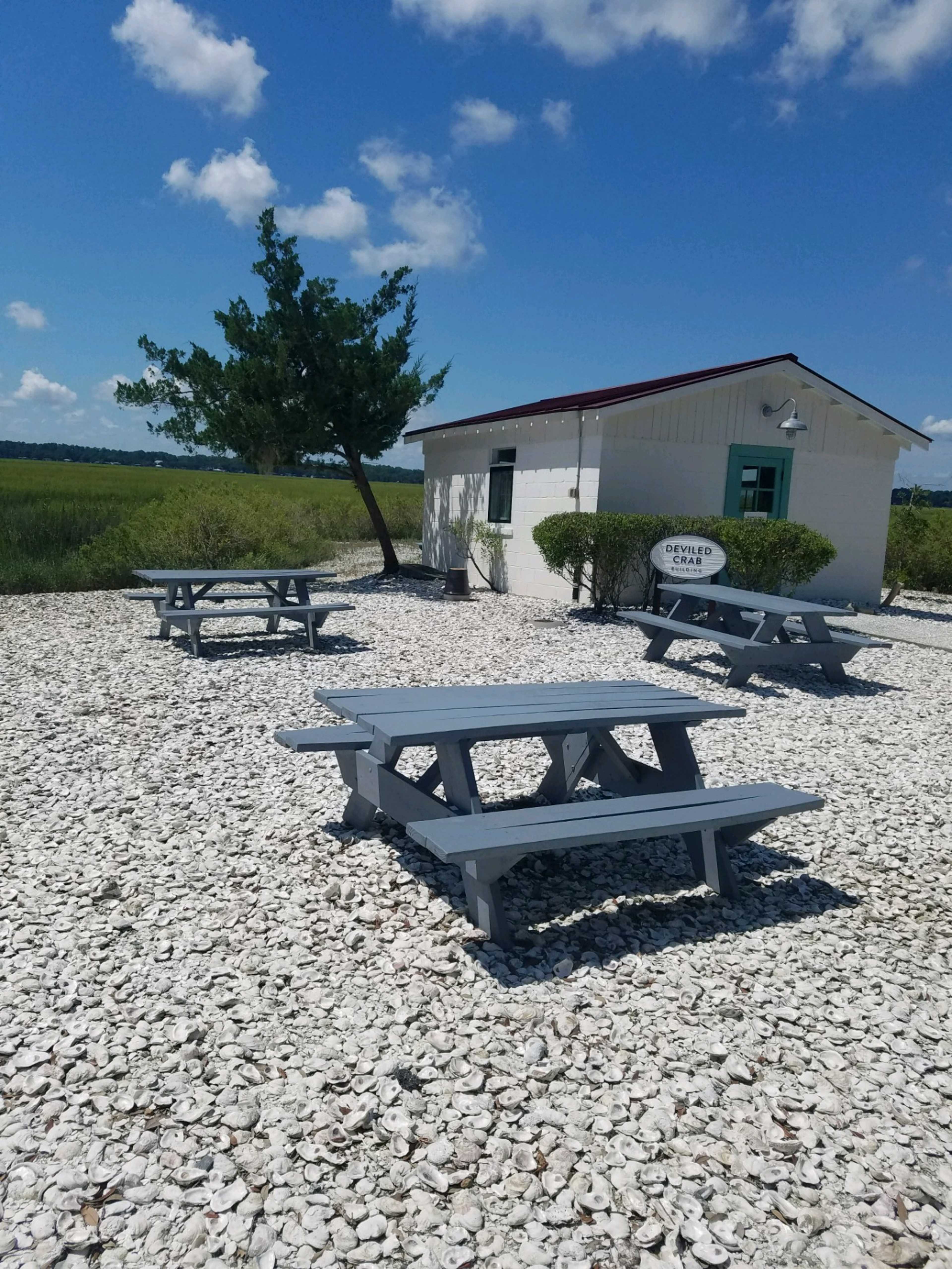 The scene depicts a small white building surrounded by picnic tables on a gravel area with greenery in the background under a blue sky.