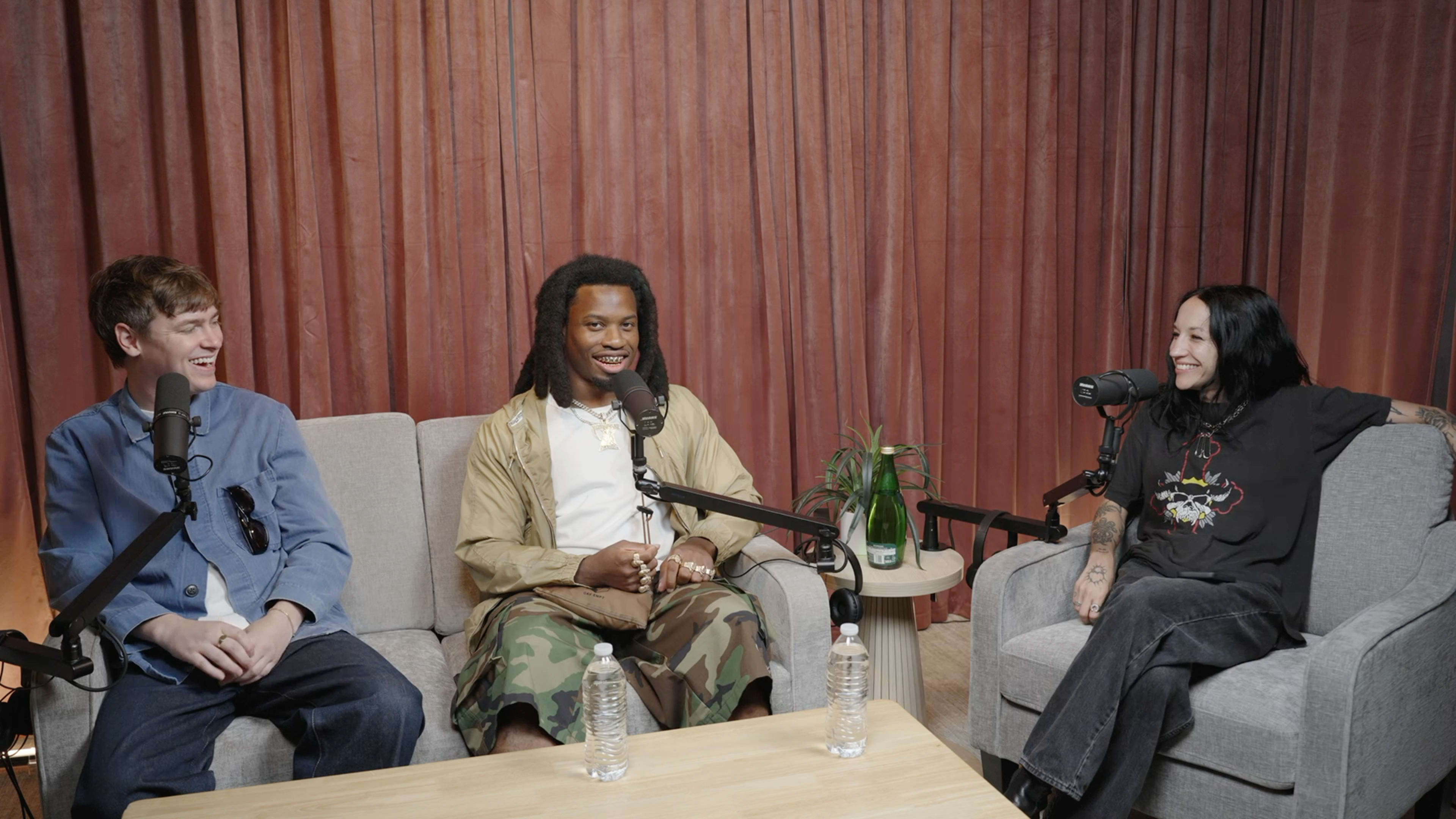 Three individuals are seated on gray sofas, engaged in conversation, with microphones positioned in front of them and a small table with water bottles in the center.