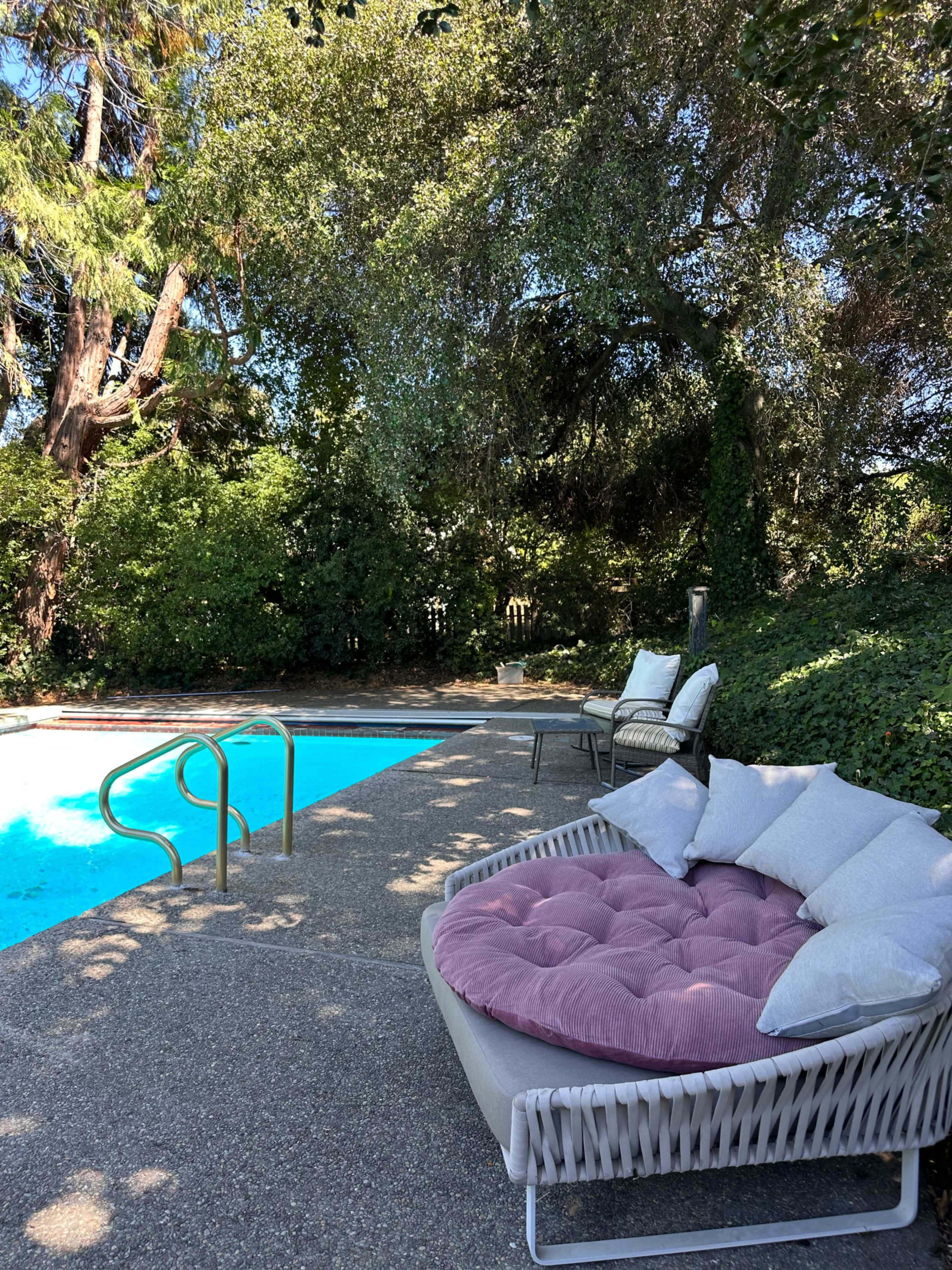 A round, pink outdoor cushion is positioned next to a swimming pool surrounded by green foliage and trees.