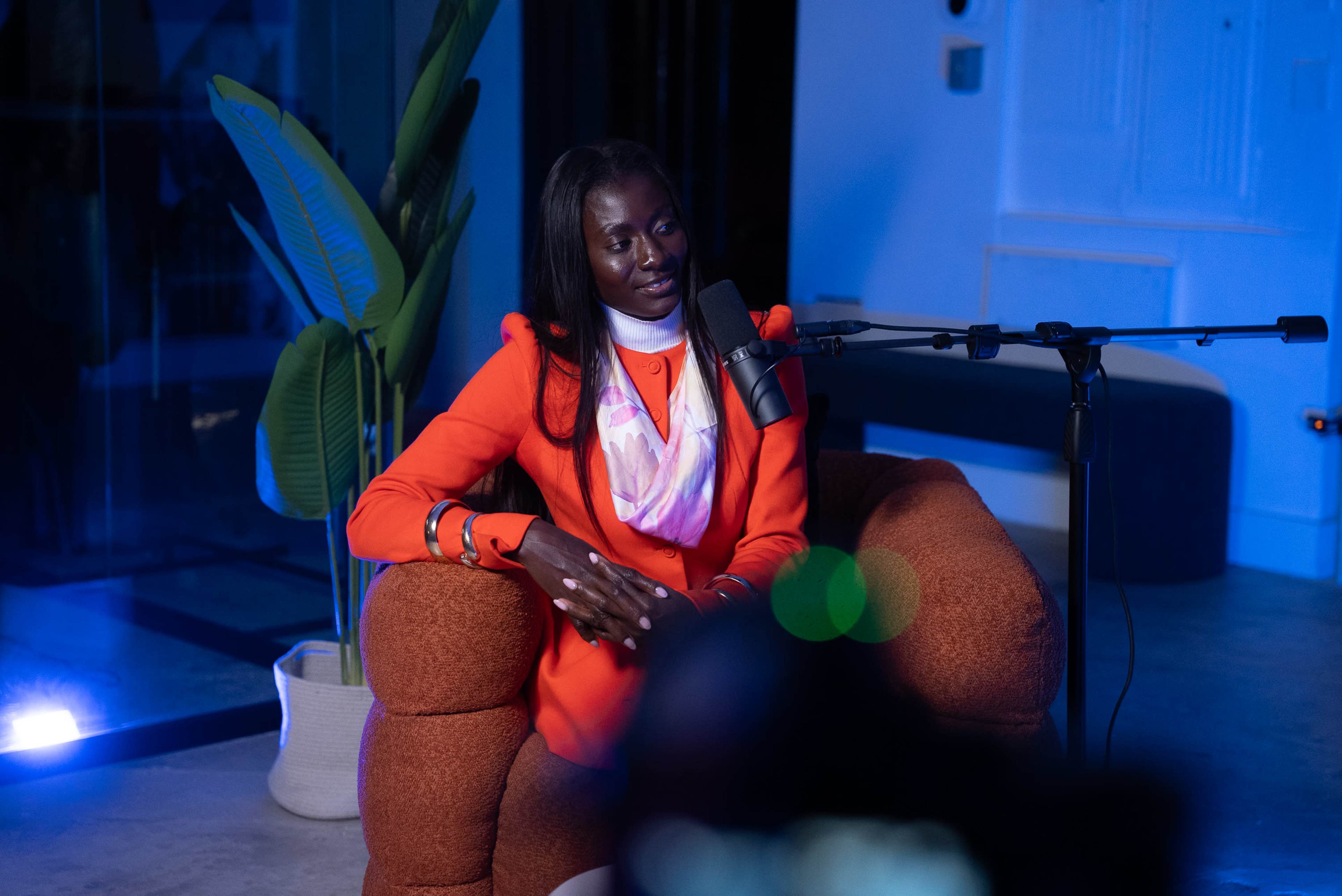 A woman in an orange blazer sits in a brown upholstered chair, speaking into a microphone during an interview in a modern setting.