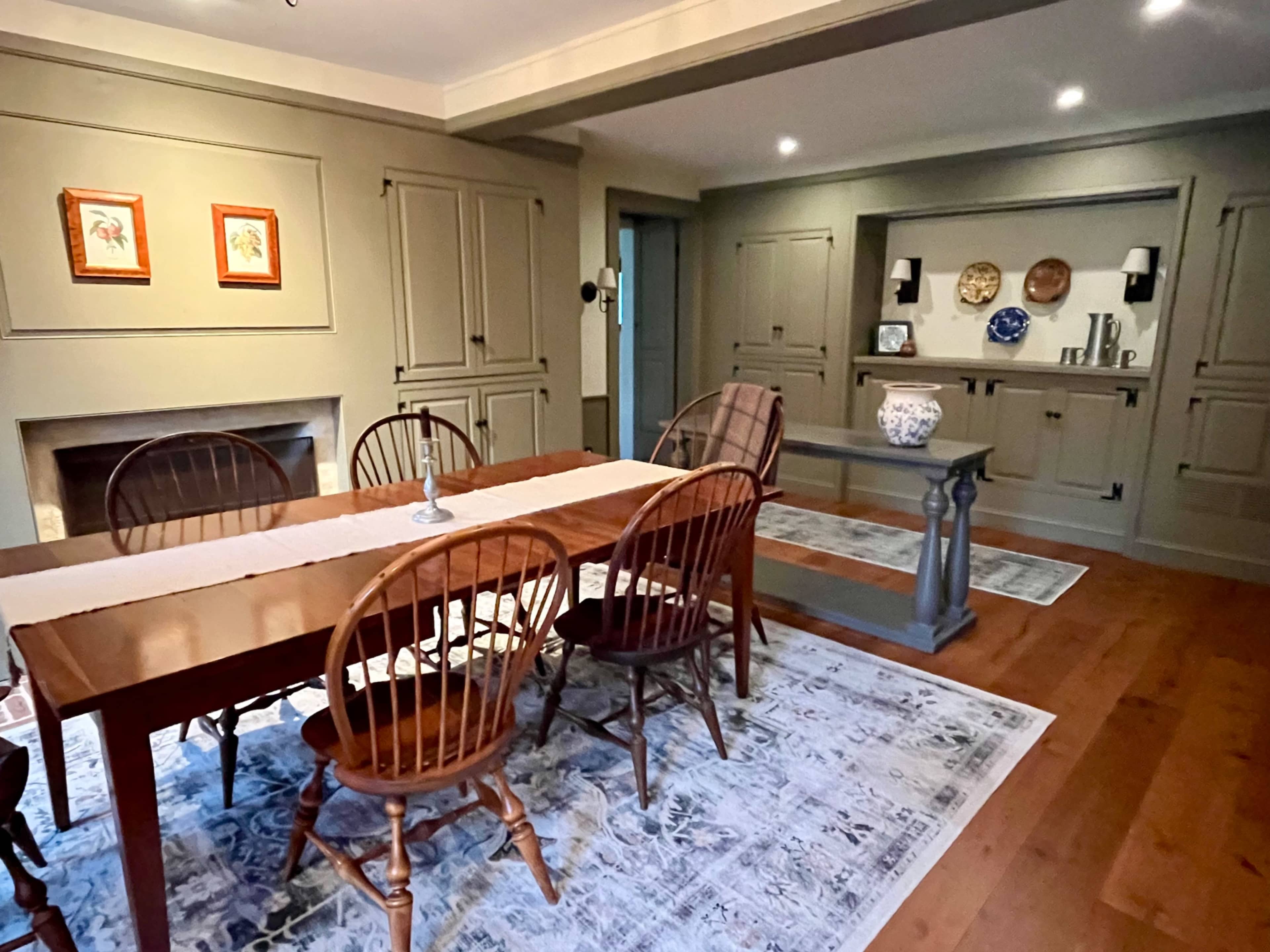 The image shows a dining room with a wooden table surrounded by chairs, a decorative area rug, and wall shelves displaying plates and other items.