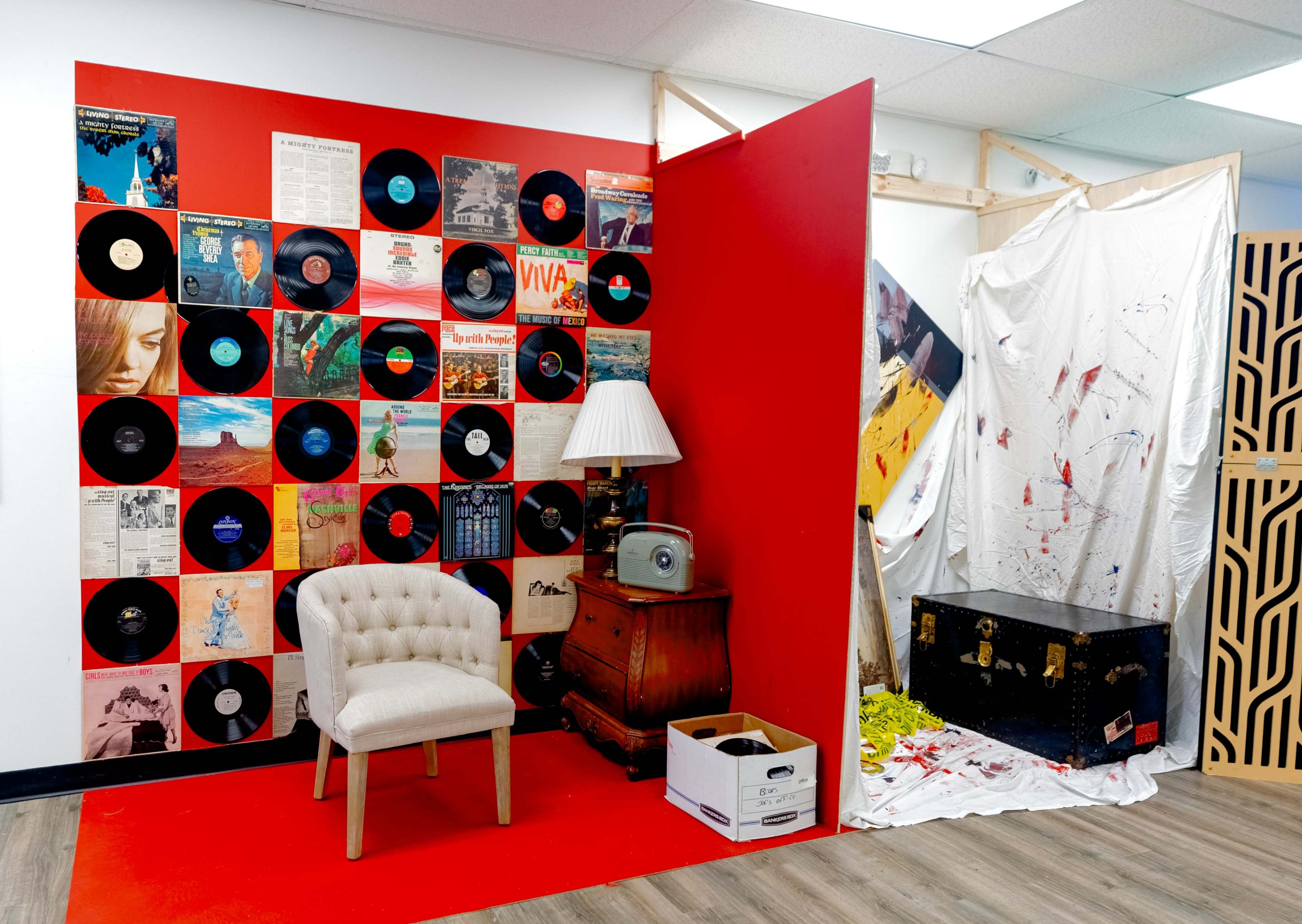 The image features a corner of a room with a red accent wall adorned with vinyl records, a vintage chair, a lamp, and a small wooden table beside a partially covered area with splattered paint.