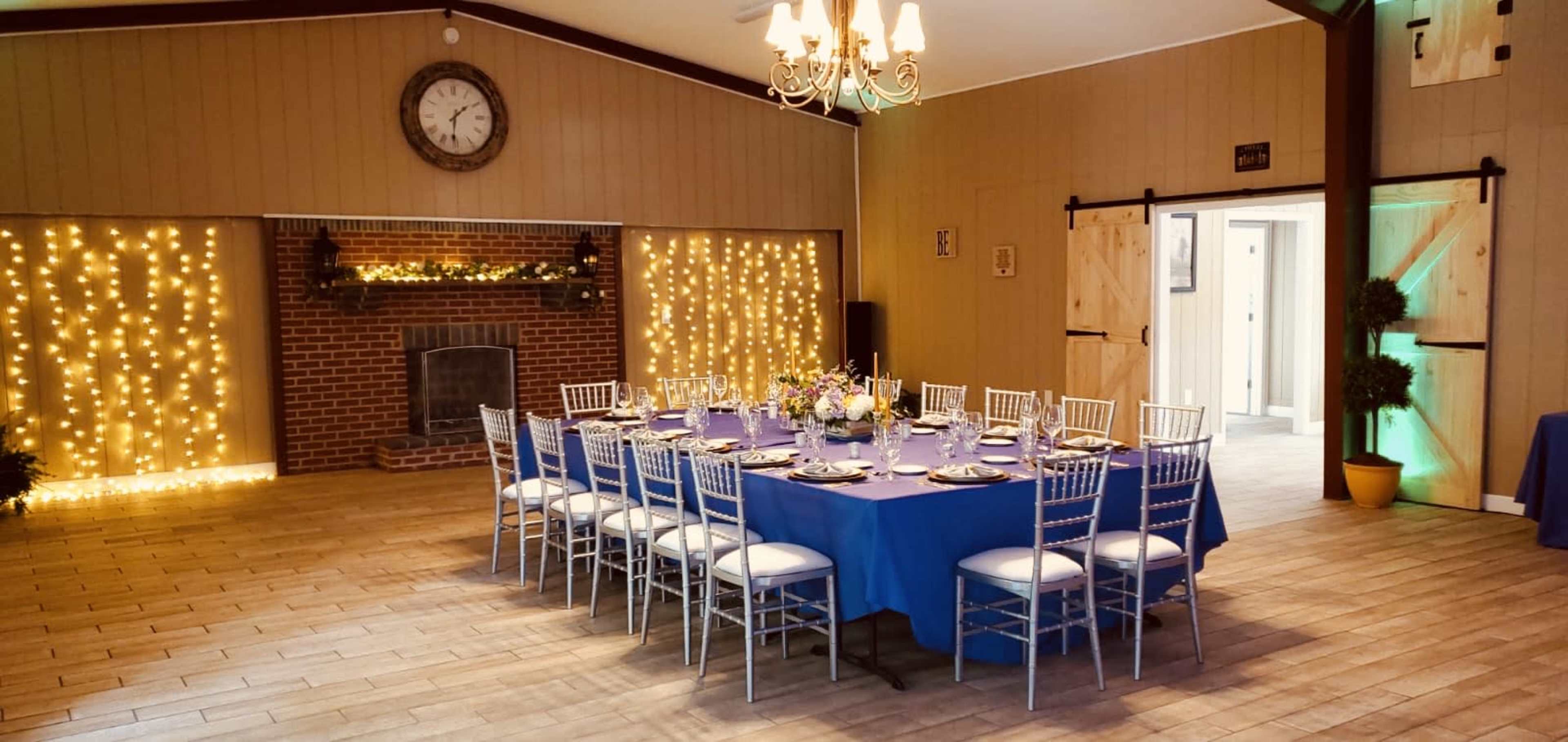 A dining area features a round table set with floral centerpieces and surrounded by silver chairs, with a brick fireplace and string lights in the background.