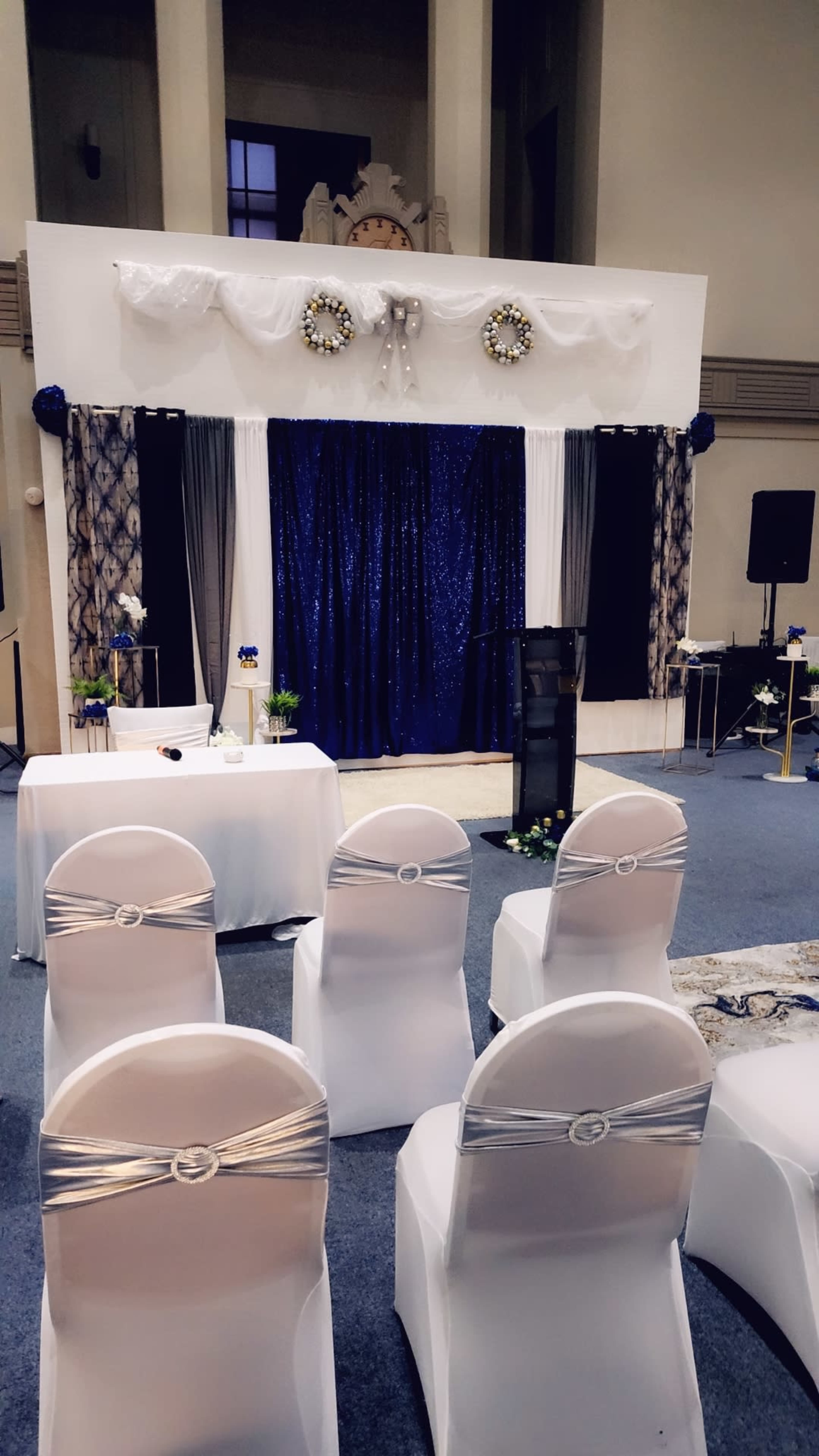 The scene shows a decorated stage with a blue backdrop, flanked by white draping and floral arrangements, in a room set up with white chairs facing the stage.
