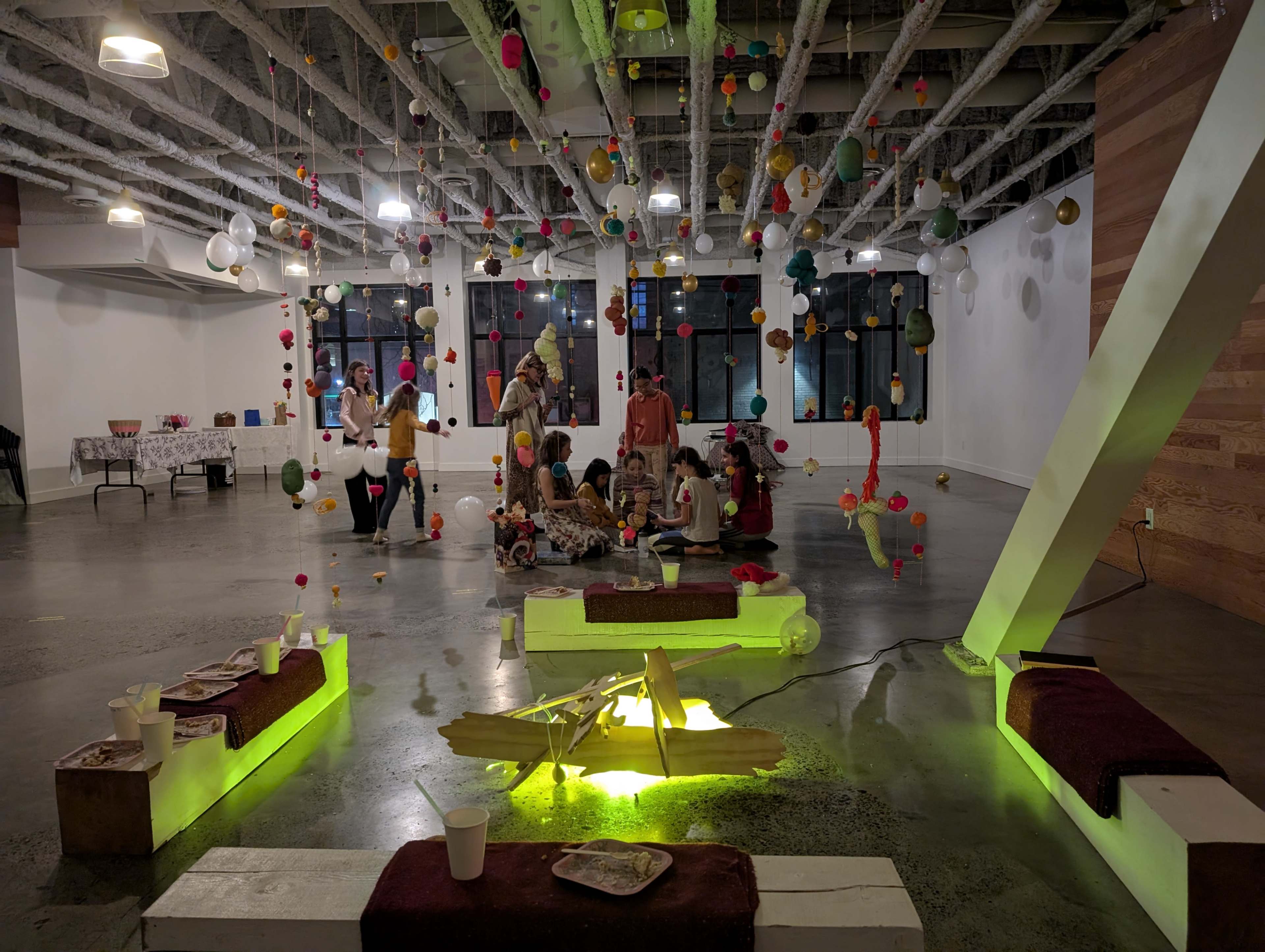 A group of people is gathered on the floor of a decorated room, surrounded by colorful hanging ornaments and illuminated by green lighting underneath low platforms.