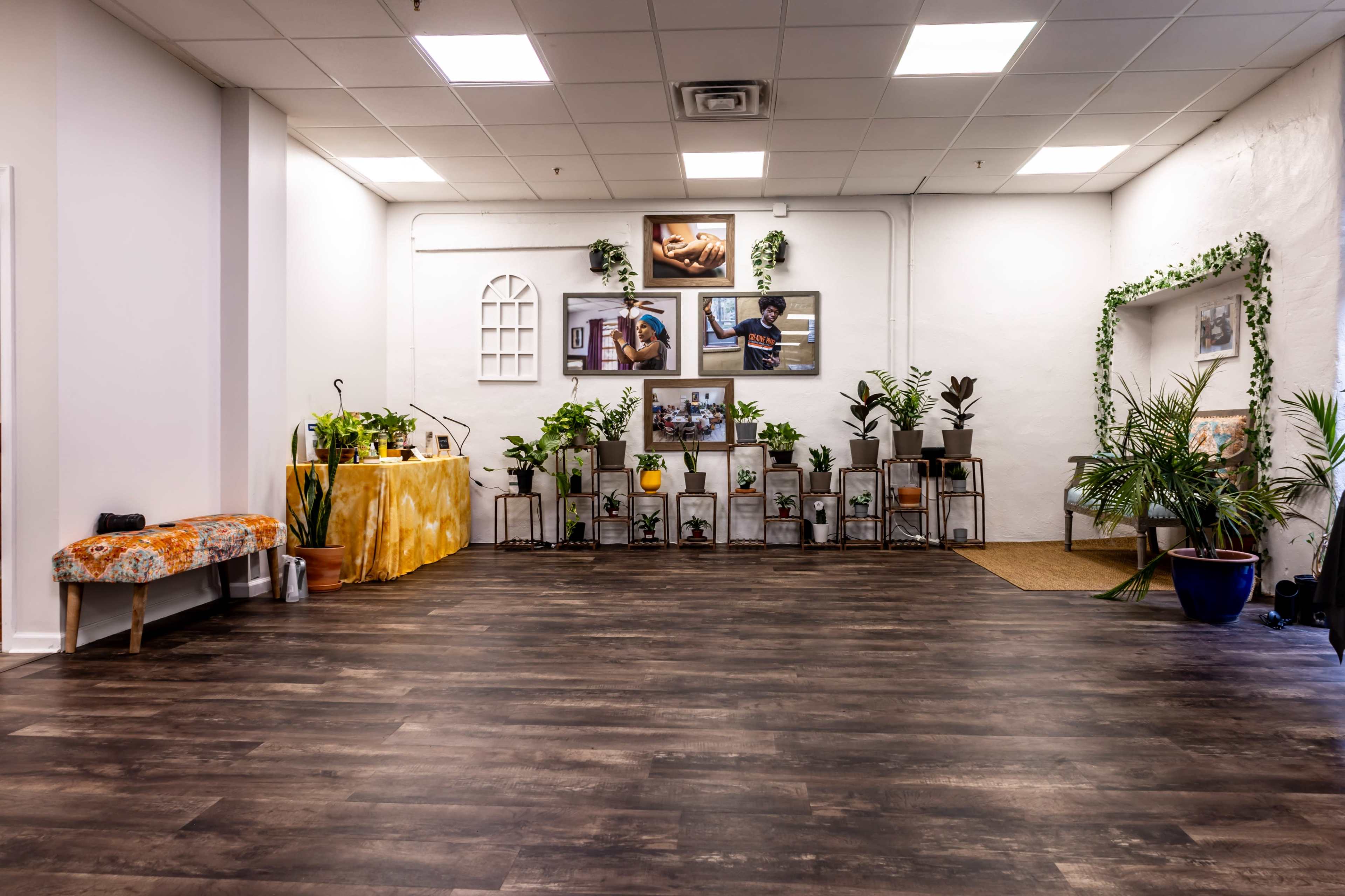 The image shows a spacious room with wooden flooring, featuring plants on shelves and walls adorned with framed pictures, along with a small table set with a colorful cloth.