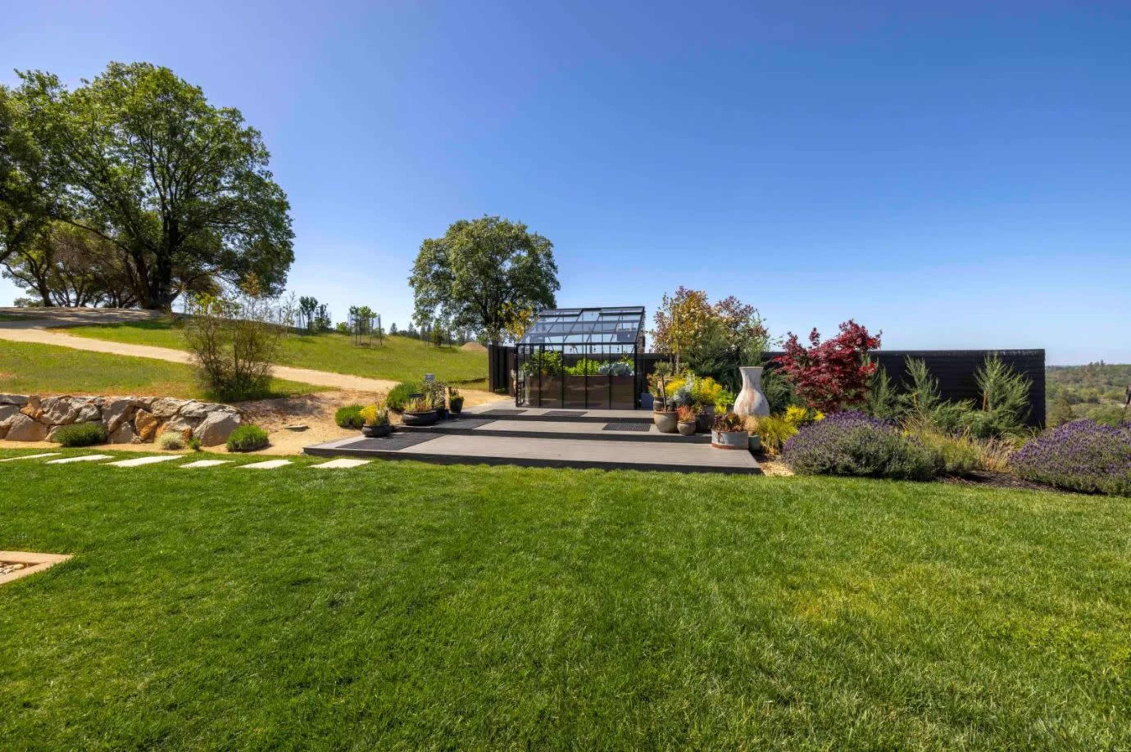 The image shows a landscaped garden with a greenhouse, surrounded by green grass and various plants under a clear blue sky.