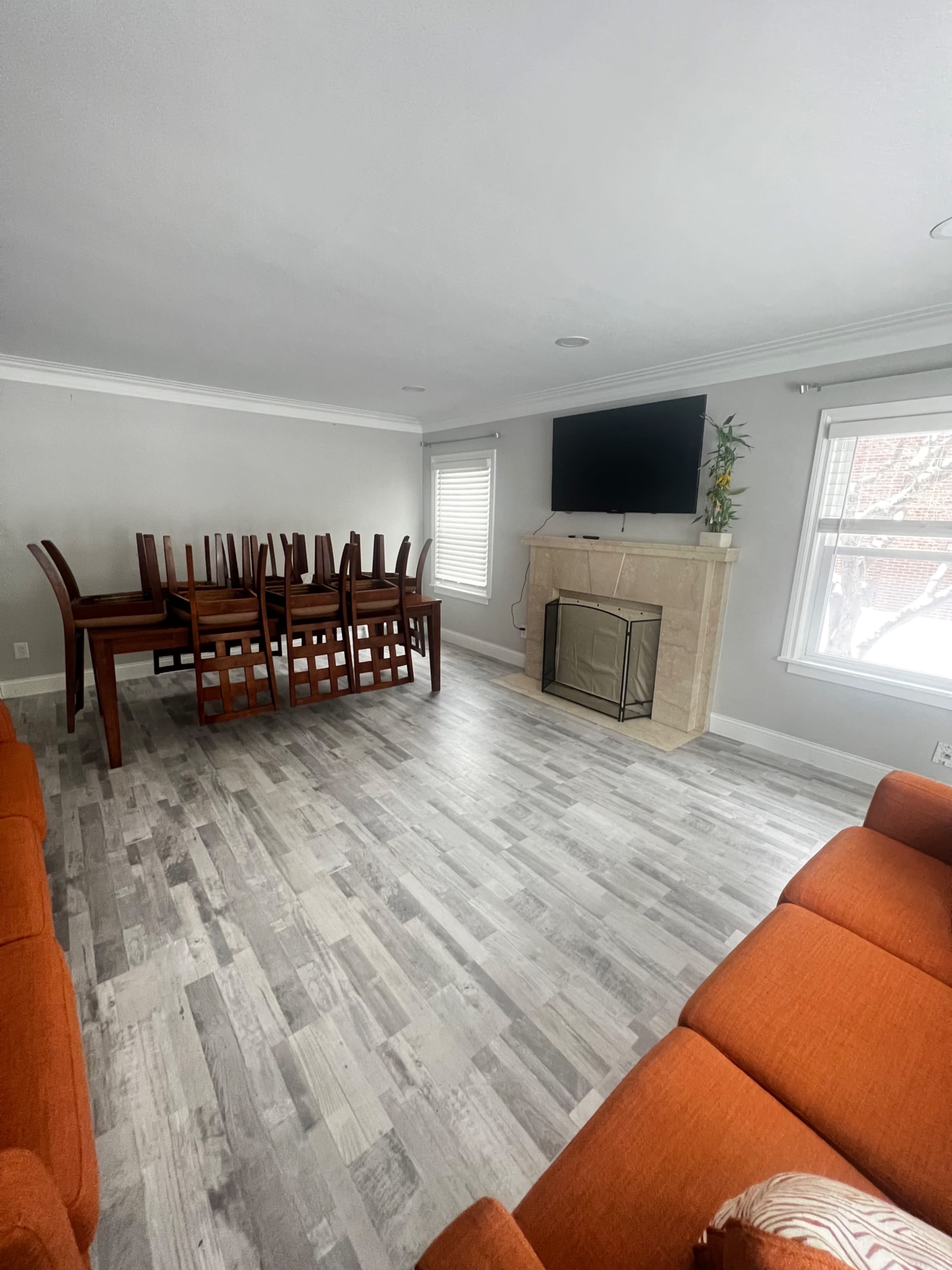 A living room features a stone fireplace, a mounted television, and a table stacked with chairs, complemented by orange sofas and a light wood floor.